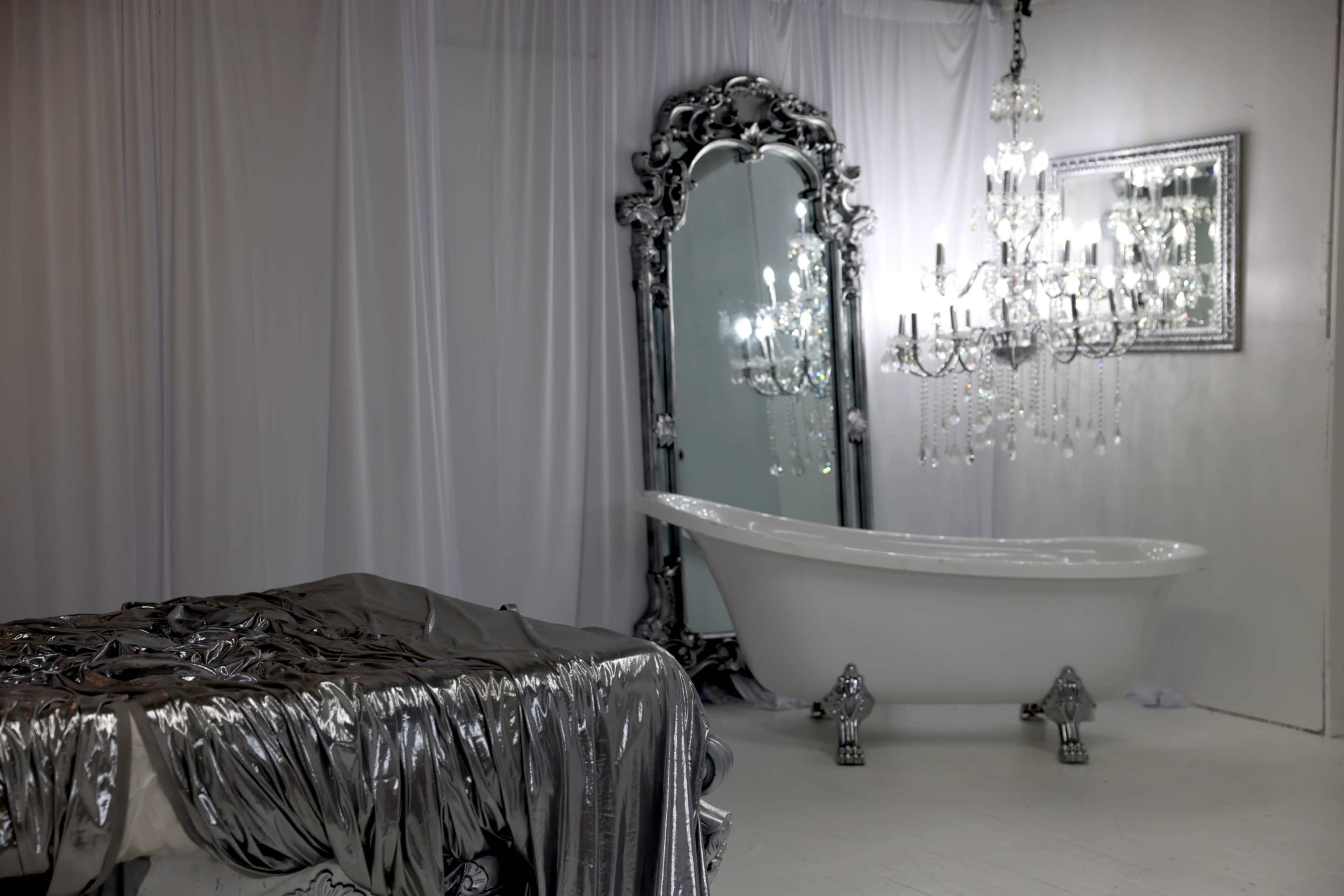 The image shows a stylish bathroom featuring a freestanding white bathtub, an ornate mirror, a crystal chandelier, and a bed draped with silver fabric.