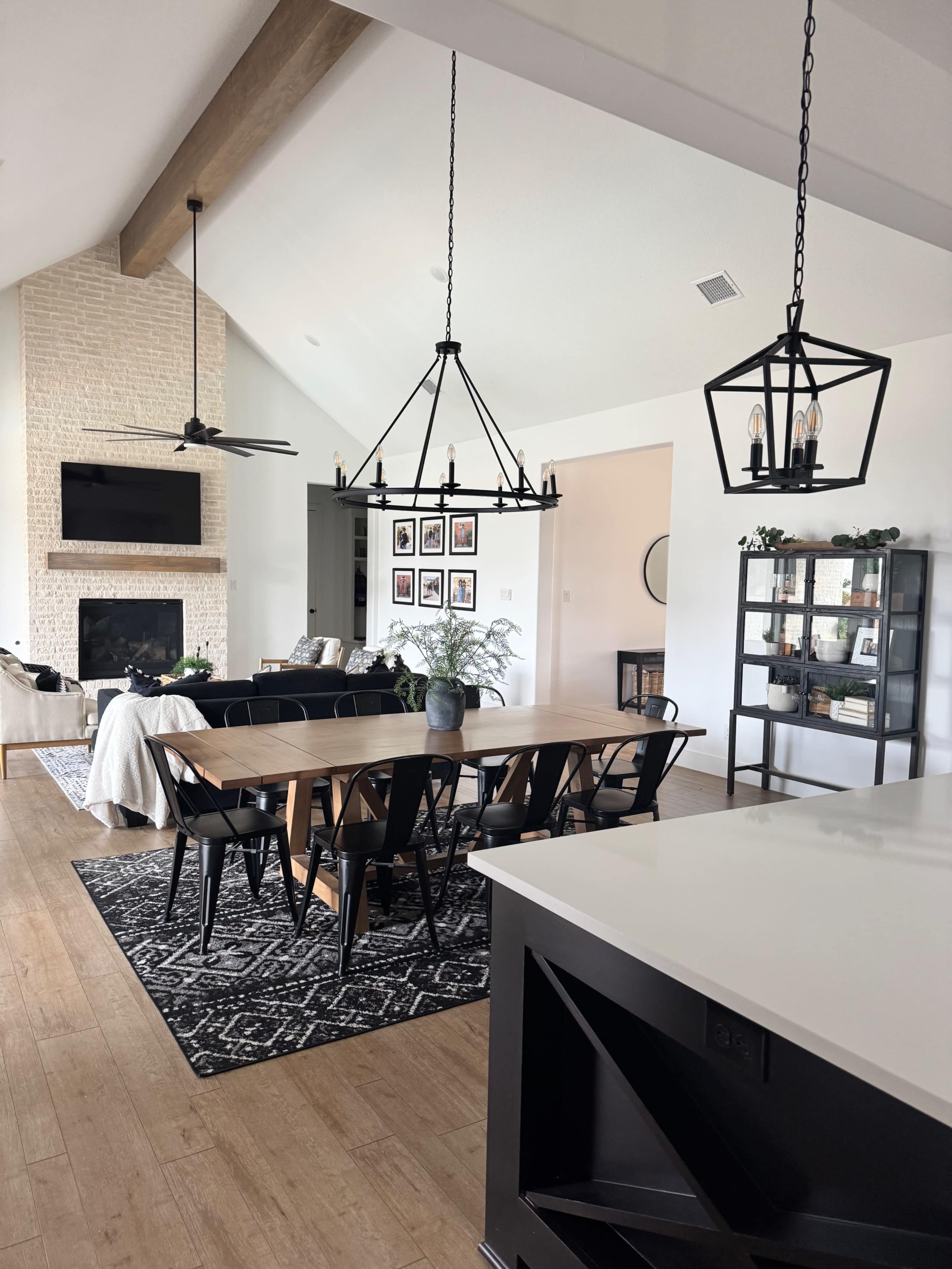 The image shows a modern dining area with a long wooden table surrounded by black chairs, adjacent to a living space with a cozy couch and a stone fireplace.