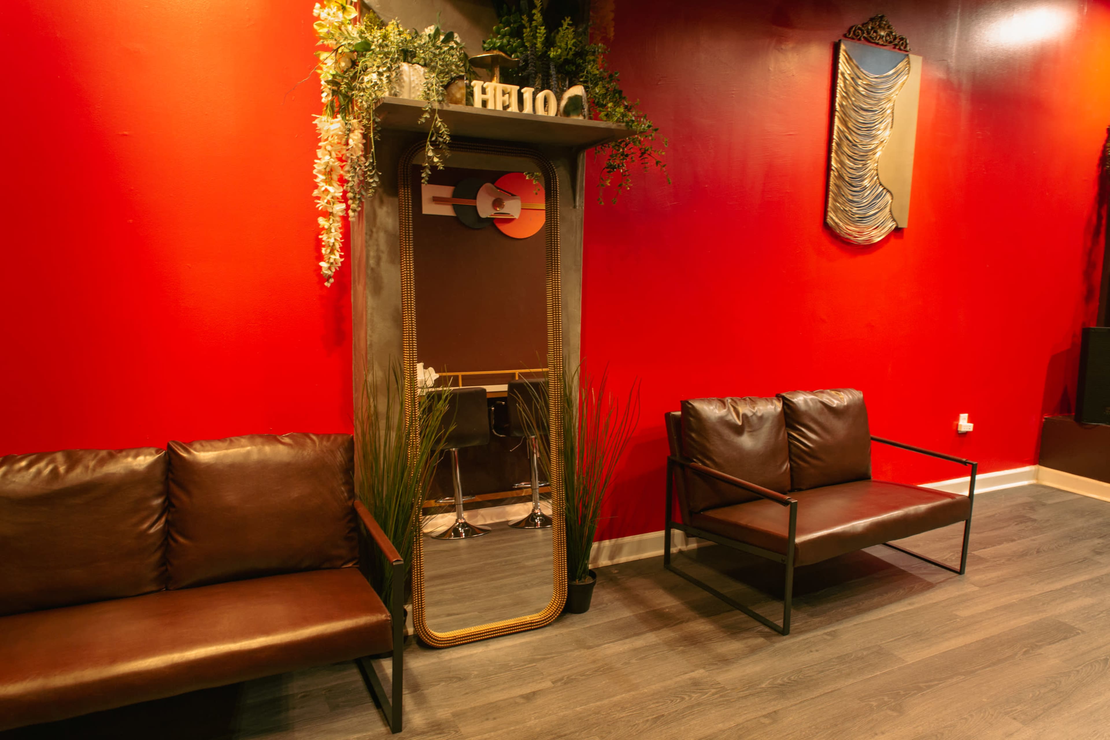 A waiting area with two brown couches, a mirror framed by greenery, and a striking red wall in the background.