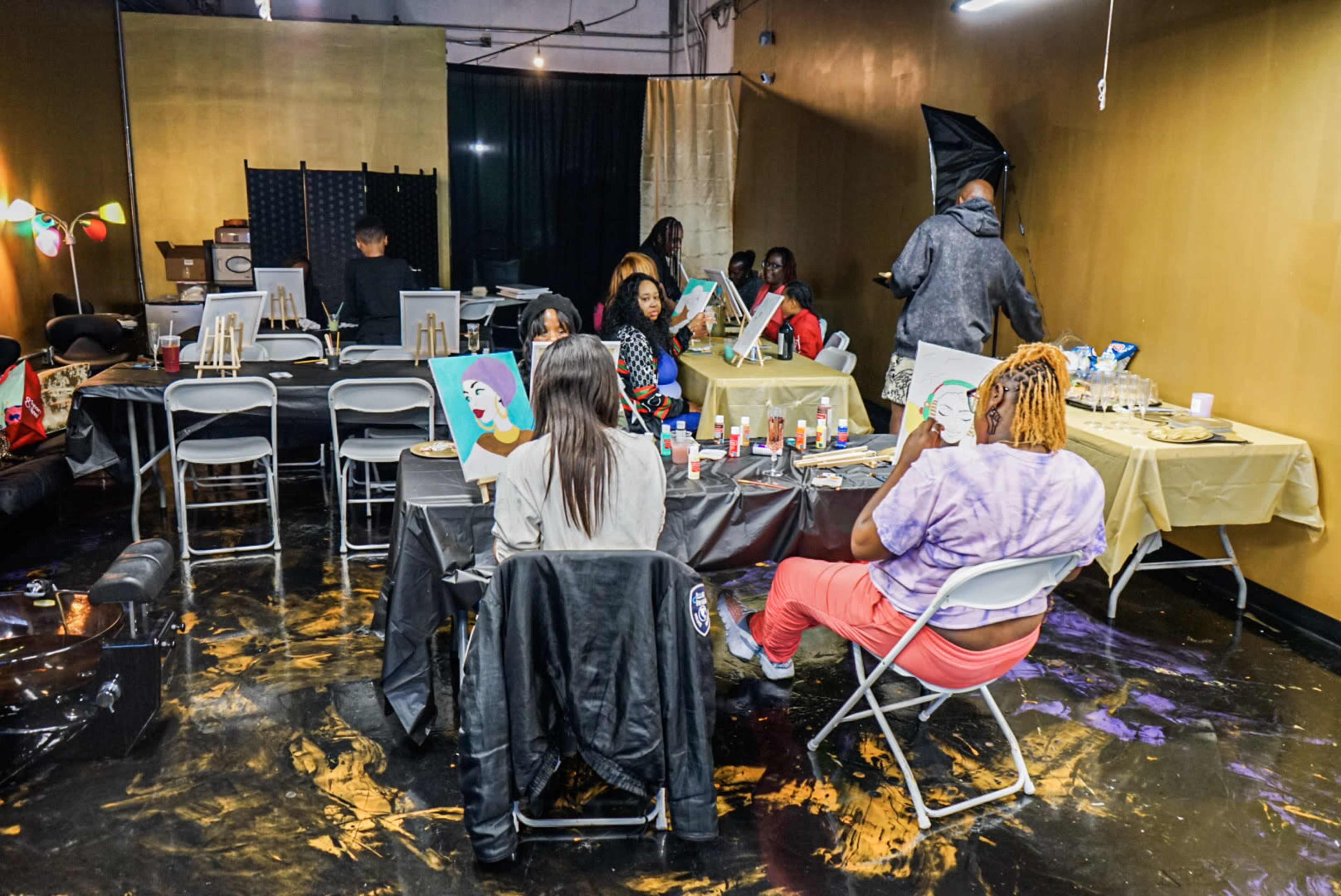 The scene shows a group of people engaged in a painting class in a dimly lit studio with gold-colored walls and various art supplies on tables.