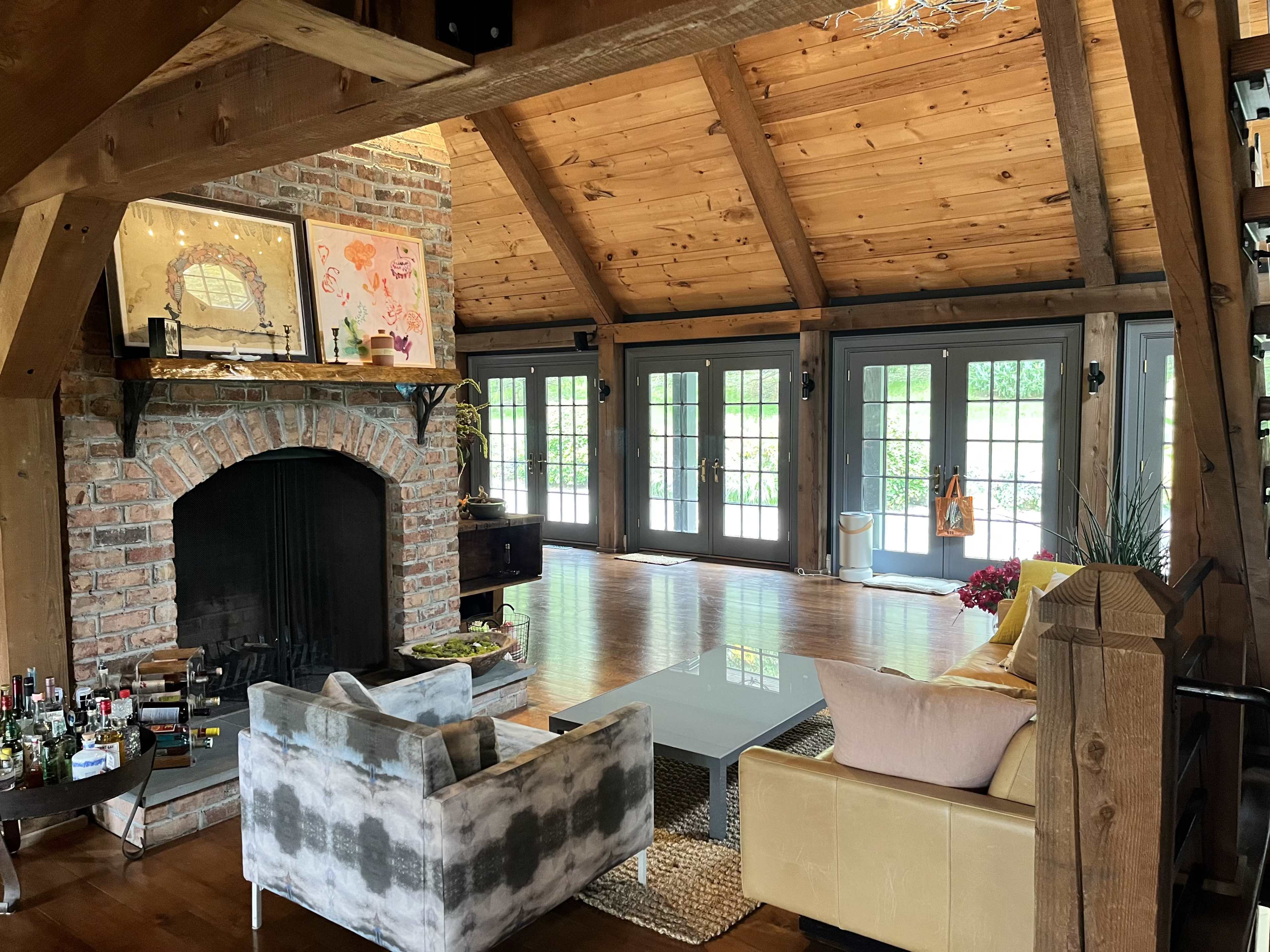 A cozy living room features a brick fireplace, wooden beams, large glass doors, and a mix of modern and traditional furniture.