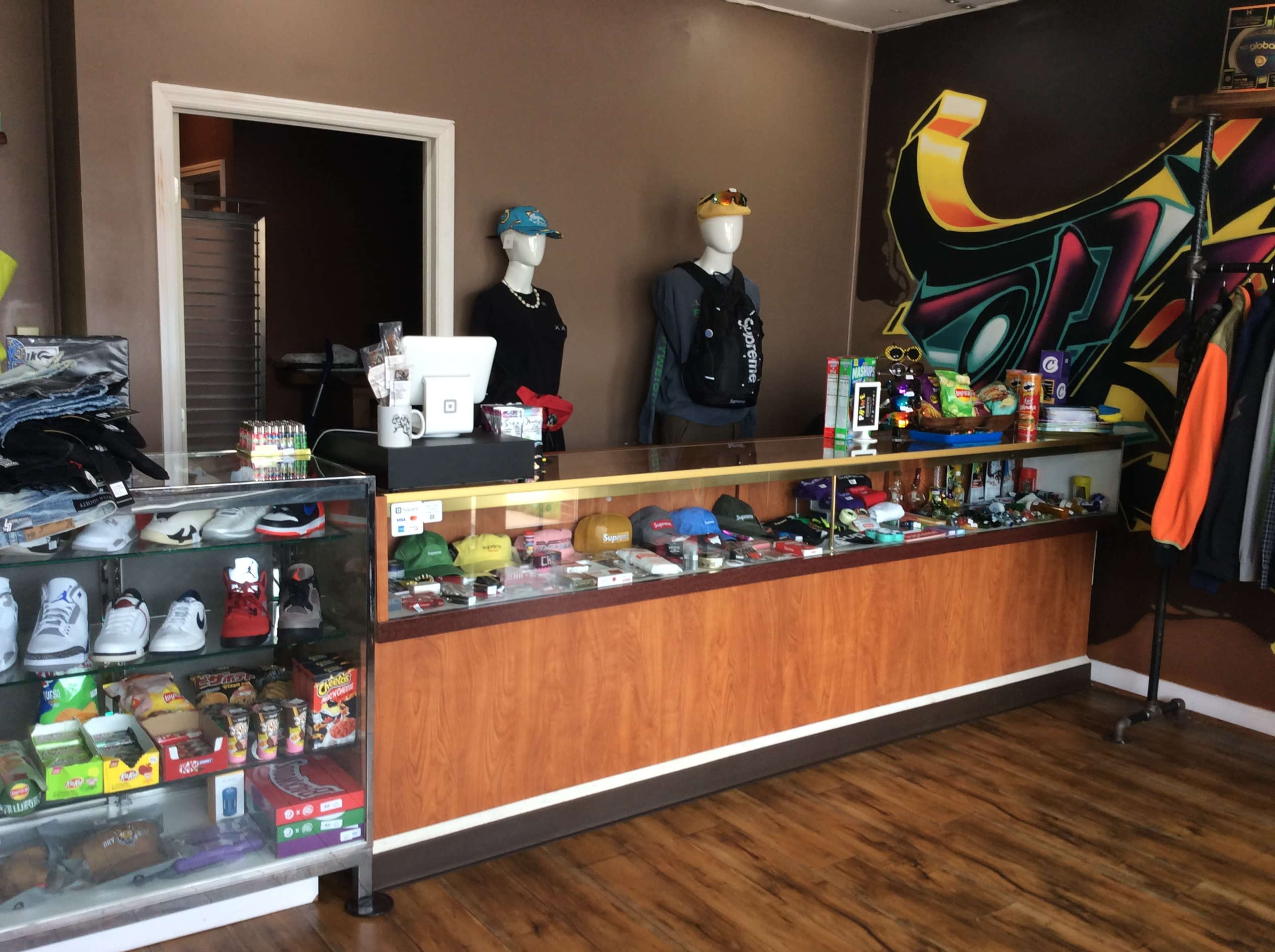 The image shows a retail store interior featuring a display counter filled with various merchandise, including clothing, accessories, and snacks, alongside mannequins dressed in apparel.
