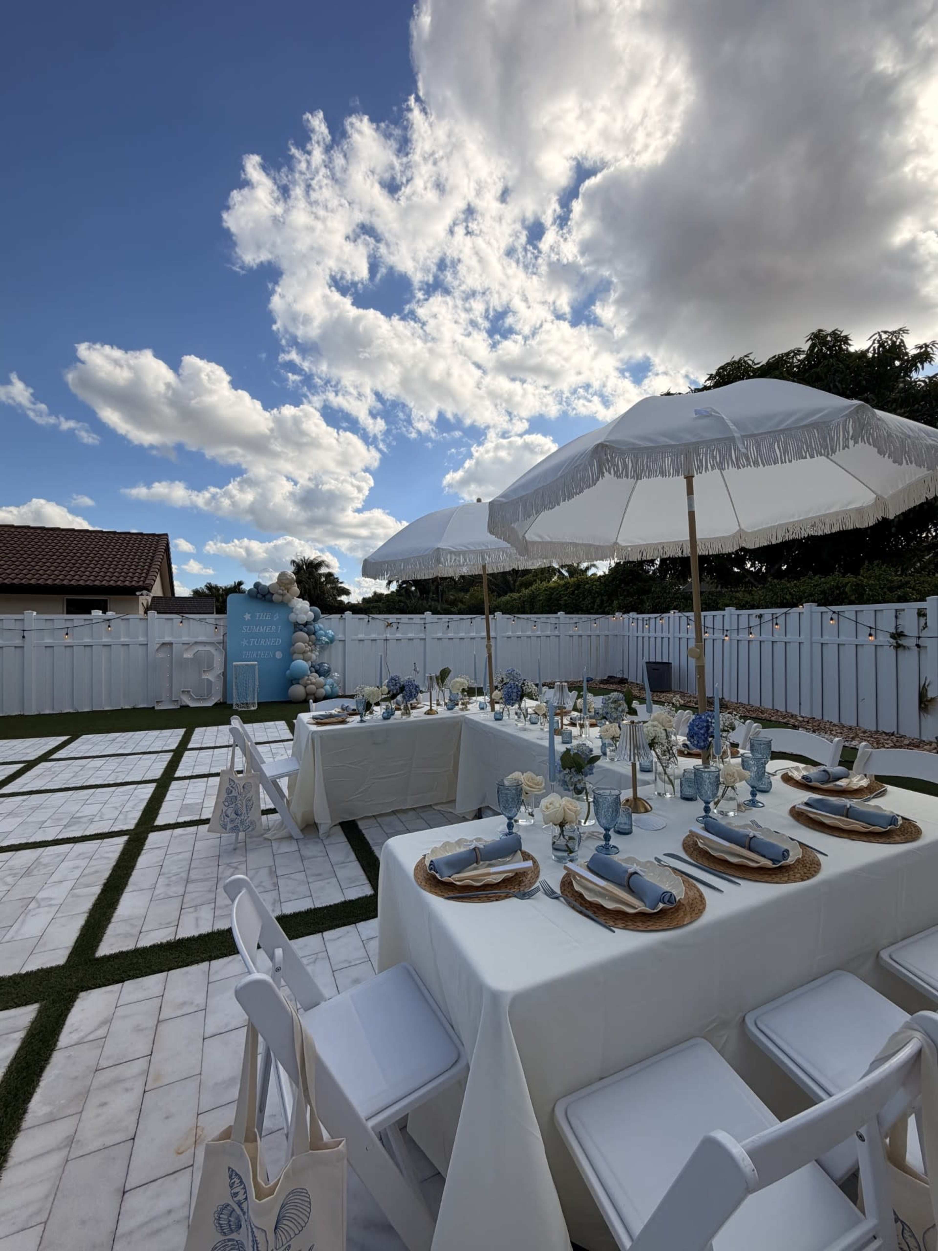 A beautifully arranged outdoor dining area features two tables set with light blue decor, surrounded by white chairs and large umbrellas, against a backdrop of fluffy clouds and a white fence.