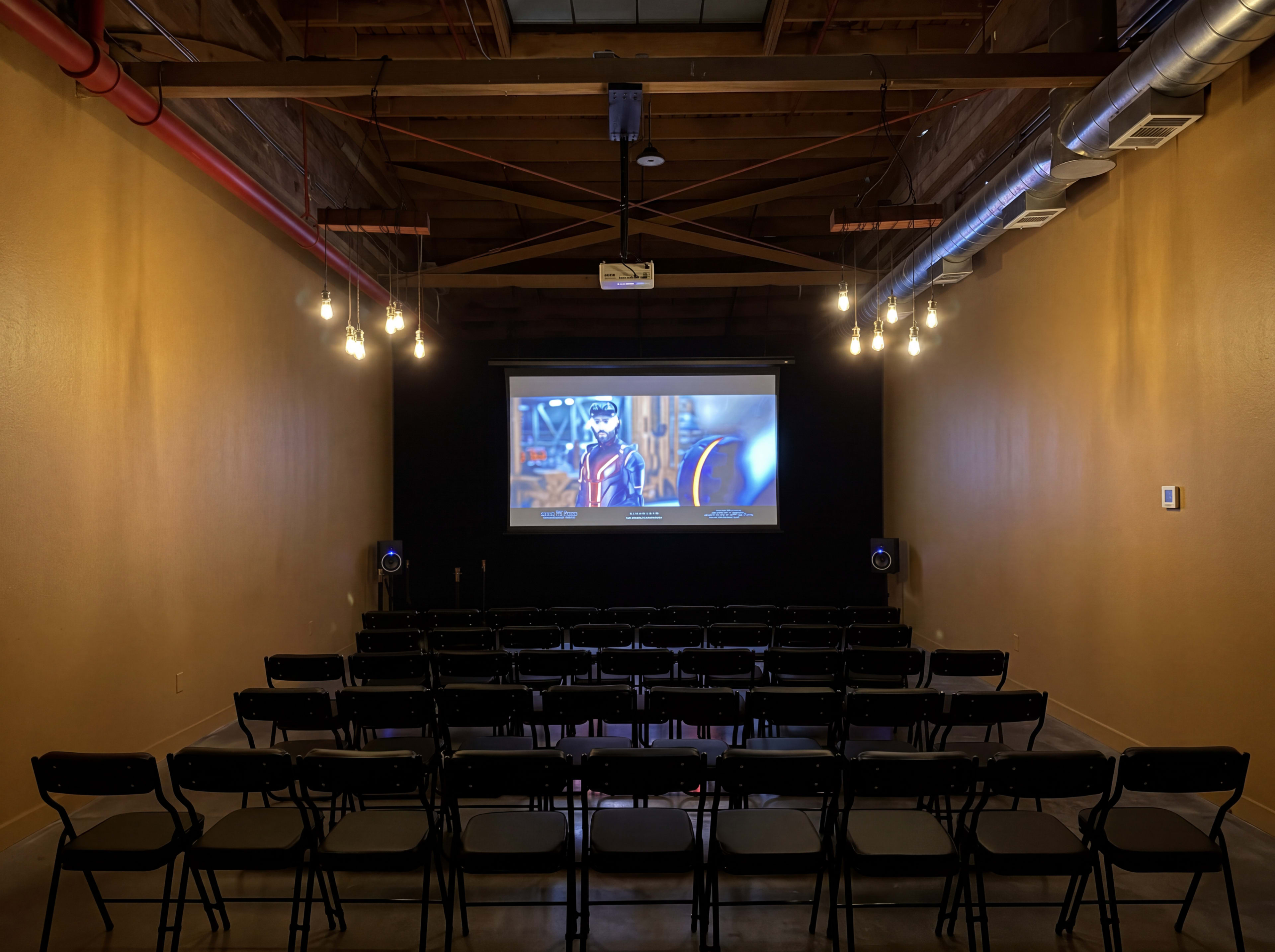 The image shows a dimly lit room with rows of black chairs facing a large projection screen displaying a film.