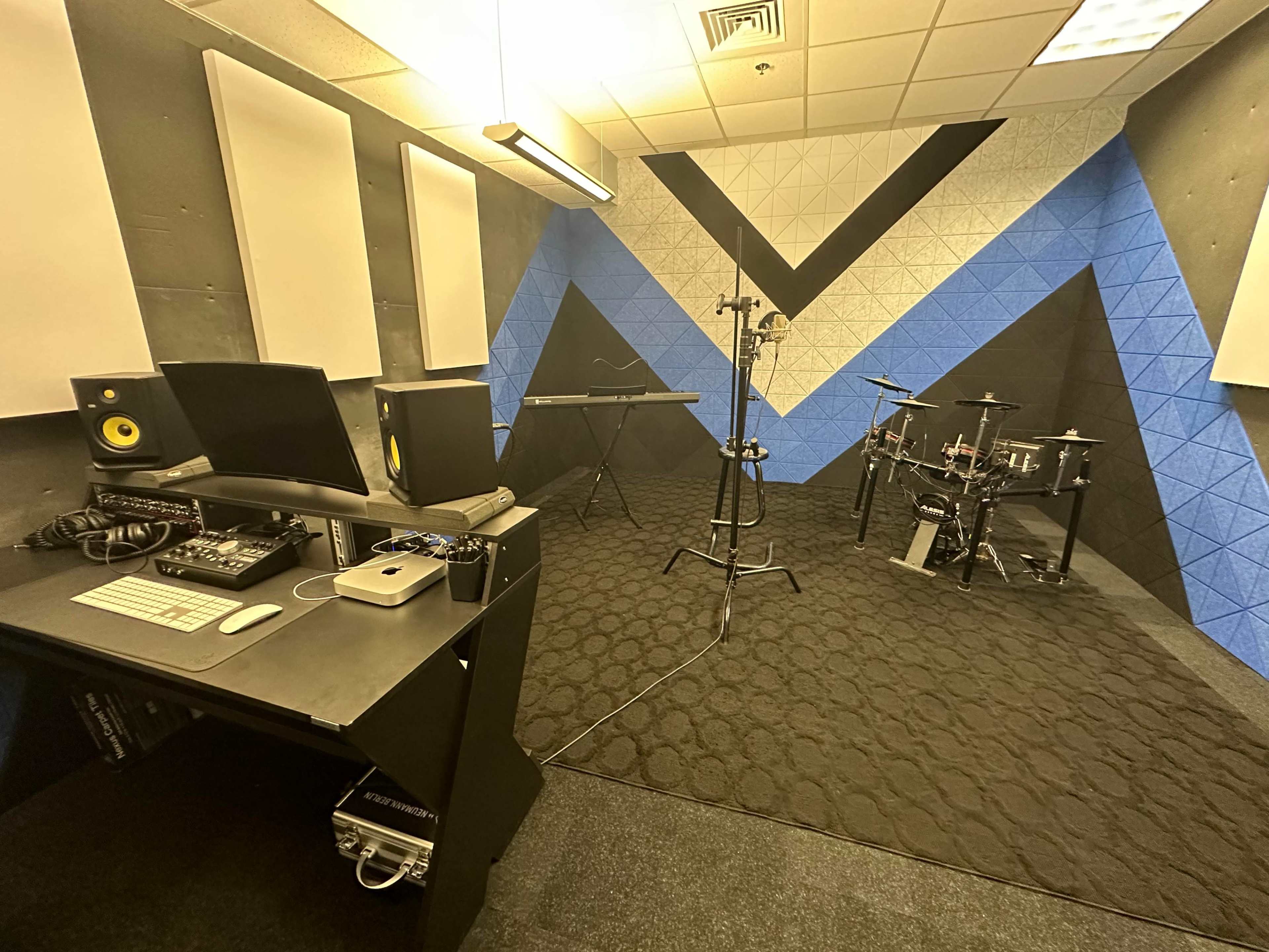 A music production room featuring a computer setup, an electronic keyboard, and a drum kit against a blue and black geometric wall.