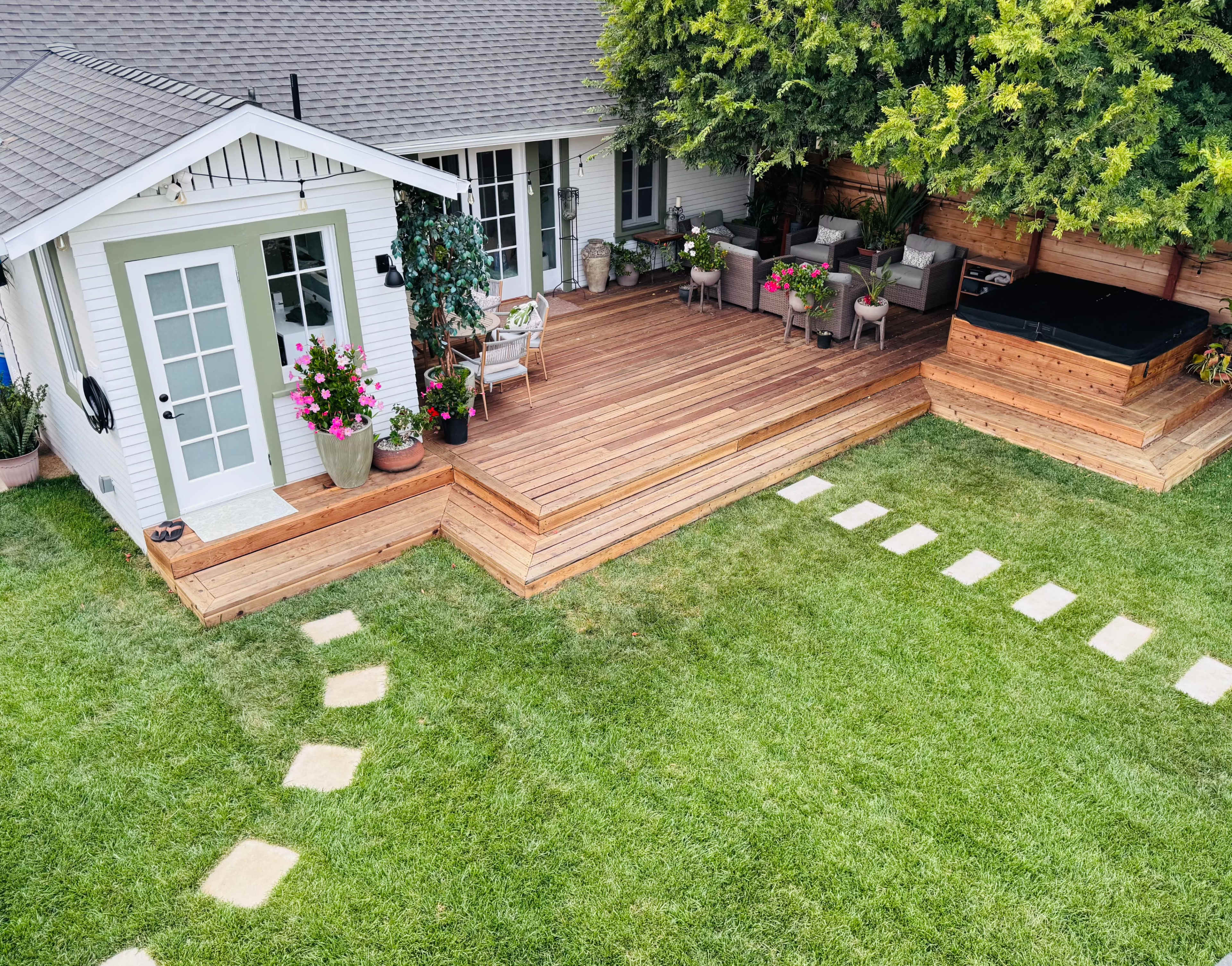 The image shows a landscaped backyard featuring a wooden deck with potted plants, seating areas, and a small shed.