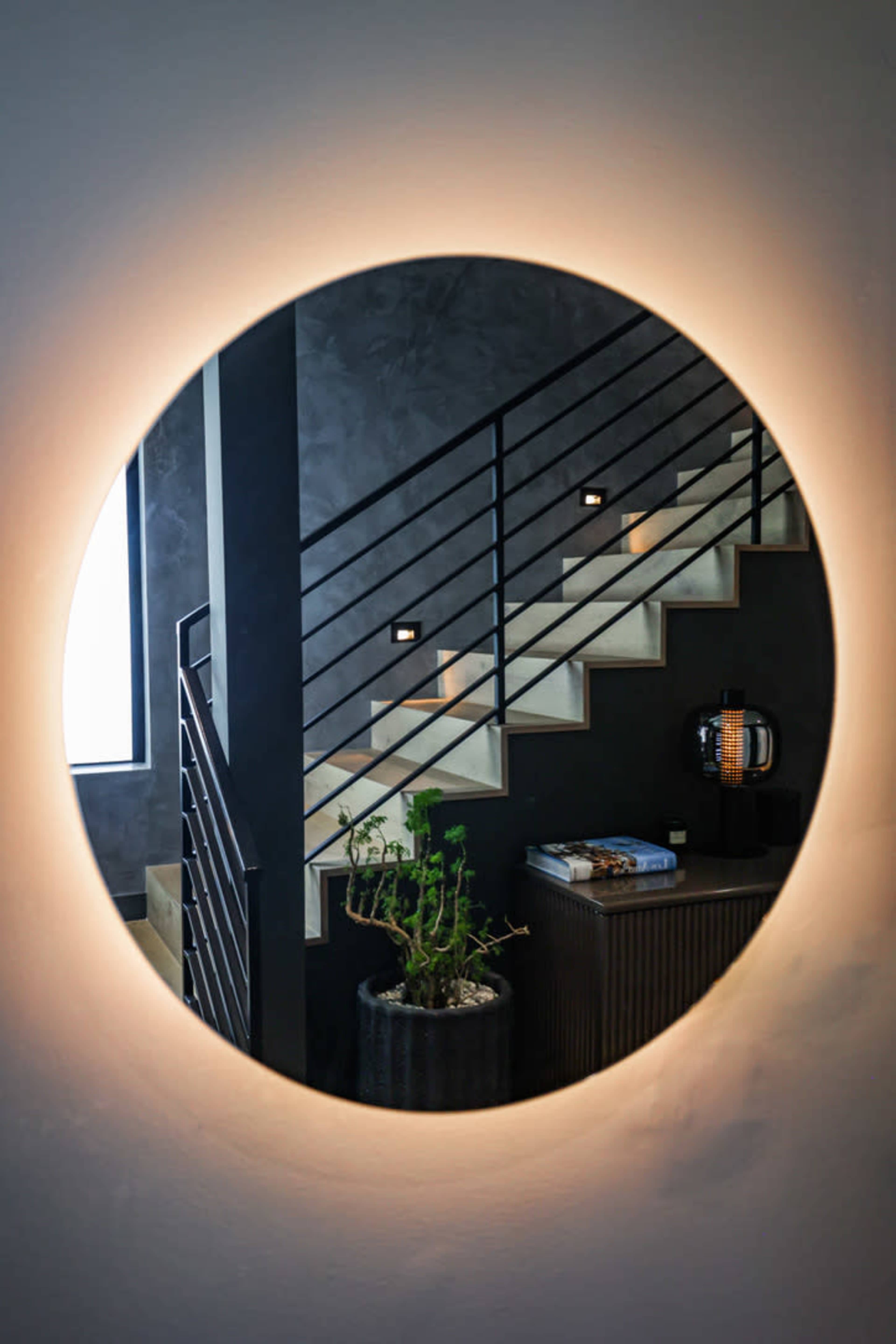A circular, illuminated mirror reflecting a modern staircase with a railing and a potted plant nearby.