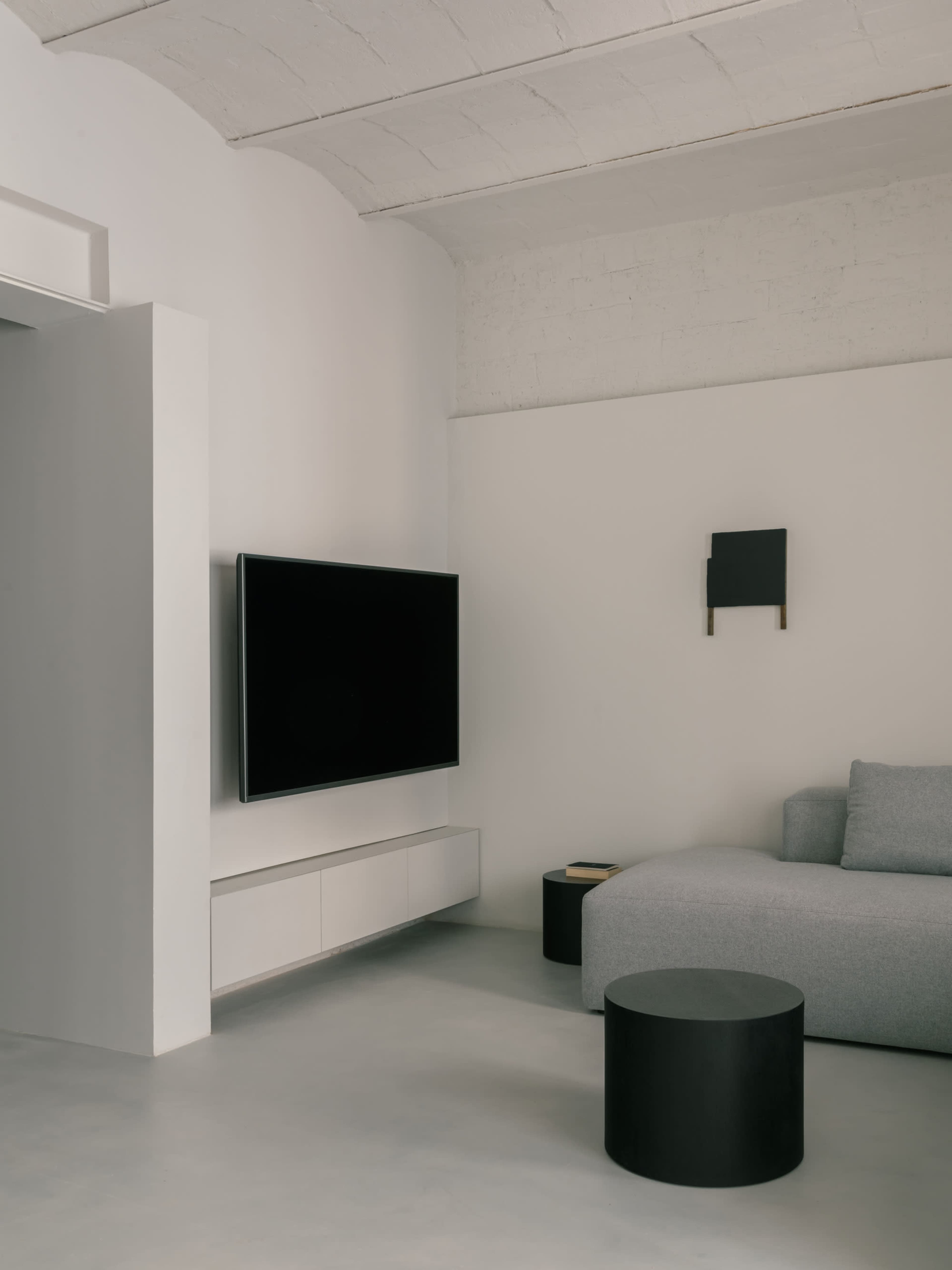 The image shows a minimalist living room featuring a gray sectional sofa, a black television mounted on the wall, and a small round black table.