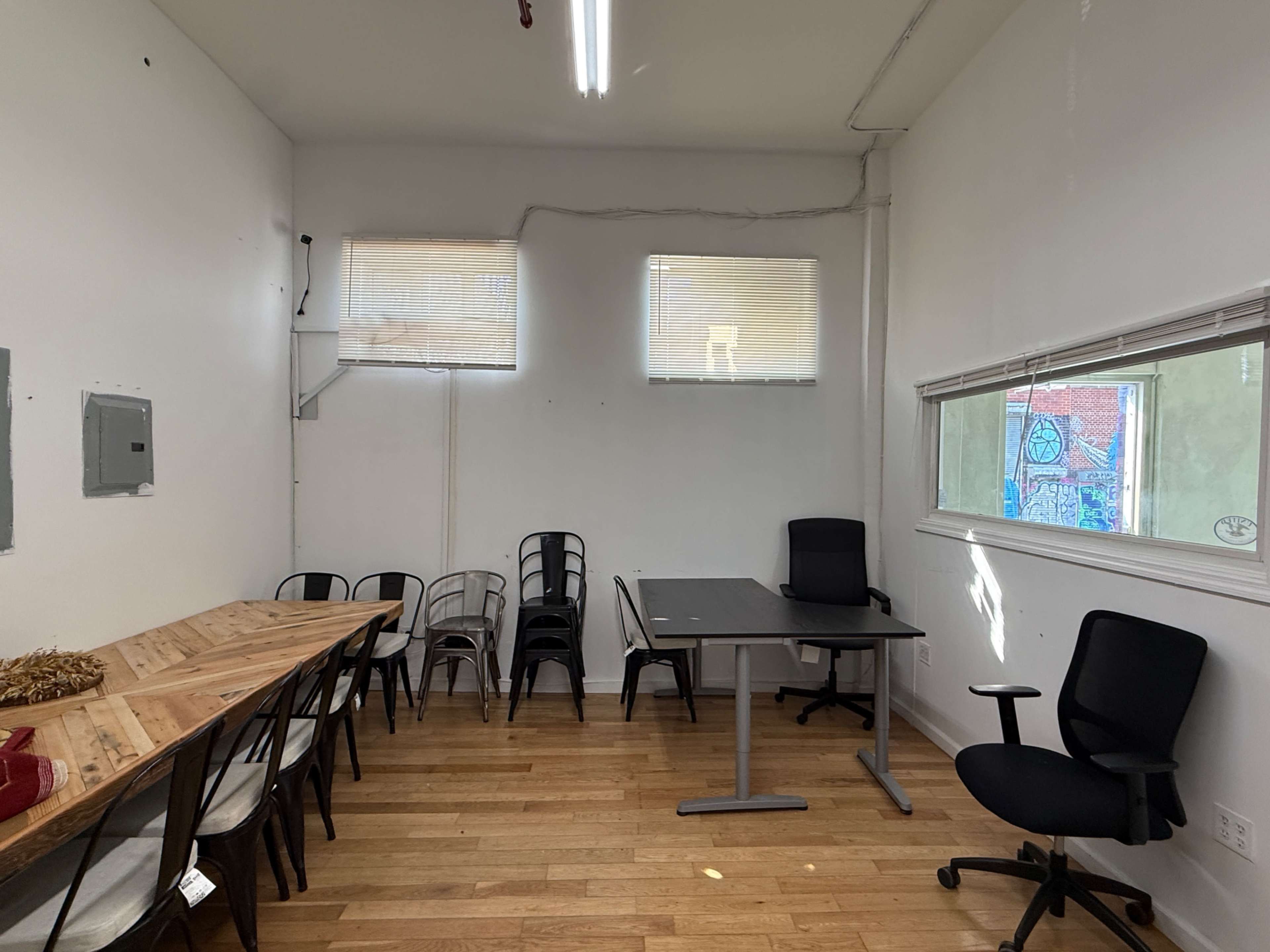 The image shows a small room with wooden flooring, two windows, a wooden table with chairs on one side, and a black table with a chair on the other side.
