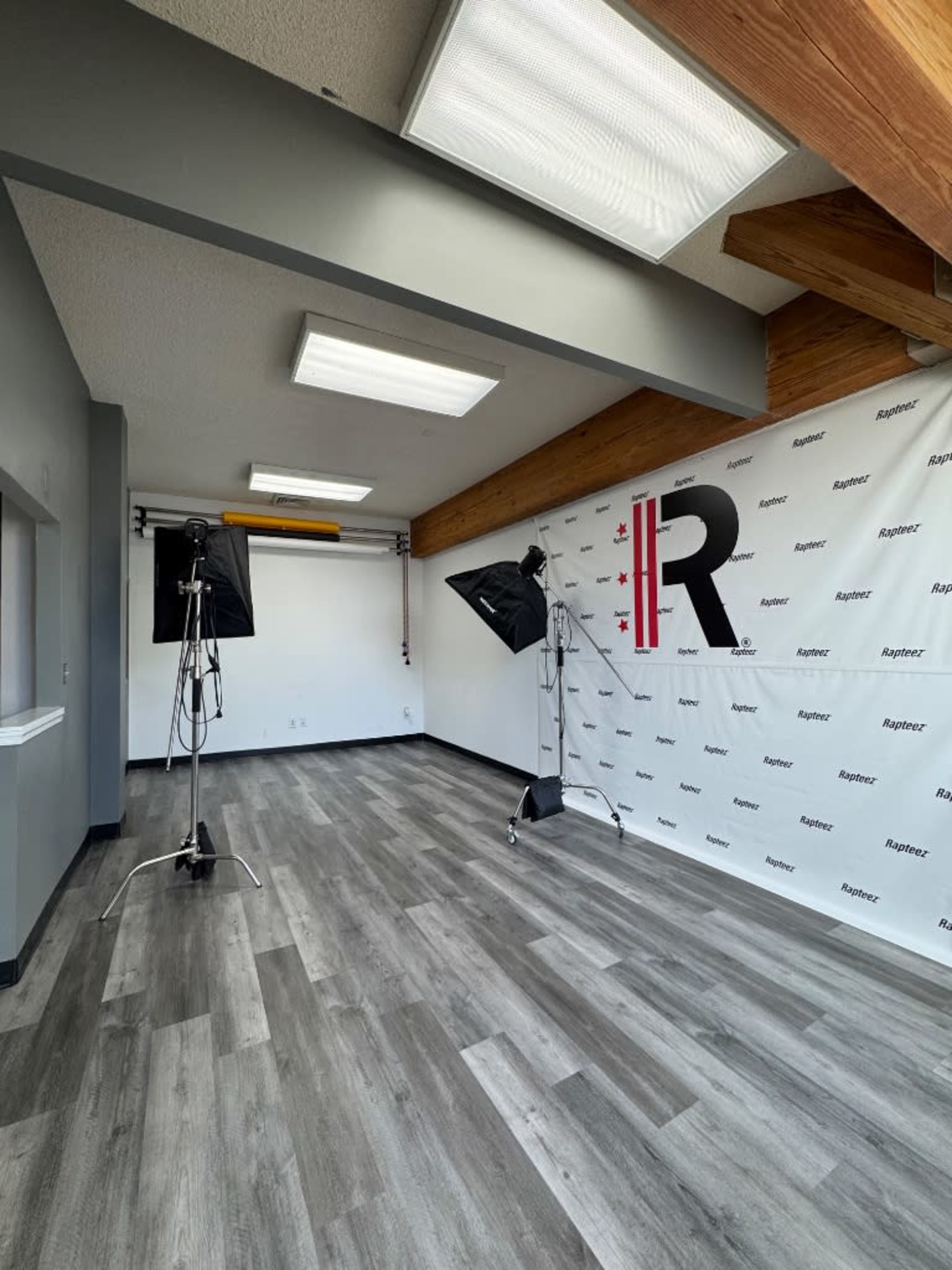 The image shows a well-lit photo studio with two lighting setups, a backdrop featuring the letter "R," and a spacious wooden-beamed ceiling.