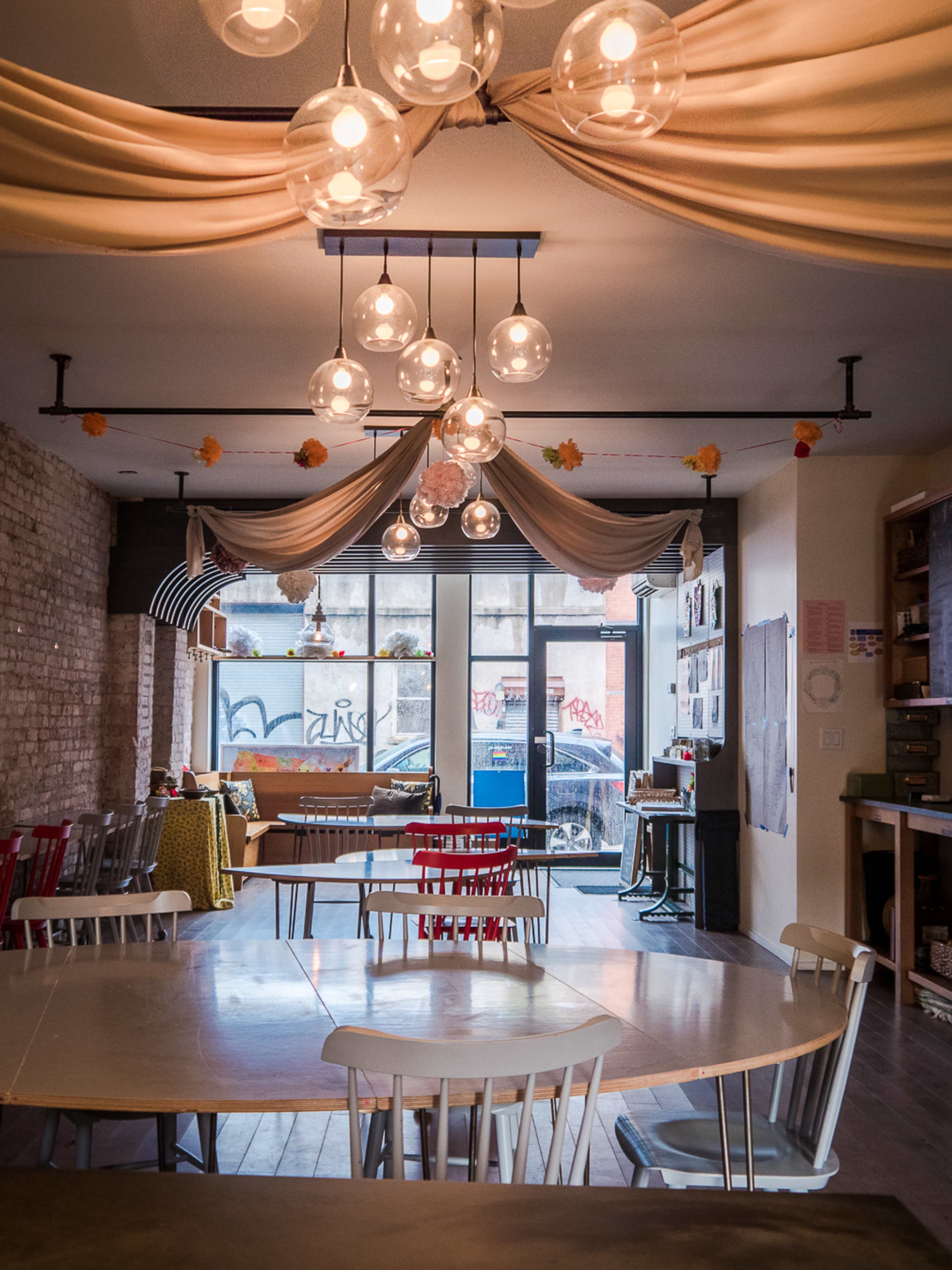 A cozy interior of a space with a large round table, various chairs, and decorative lighting, featuring windows overlooking a street with graffiti.