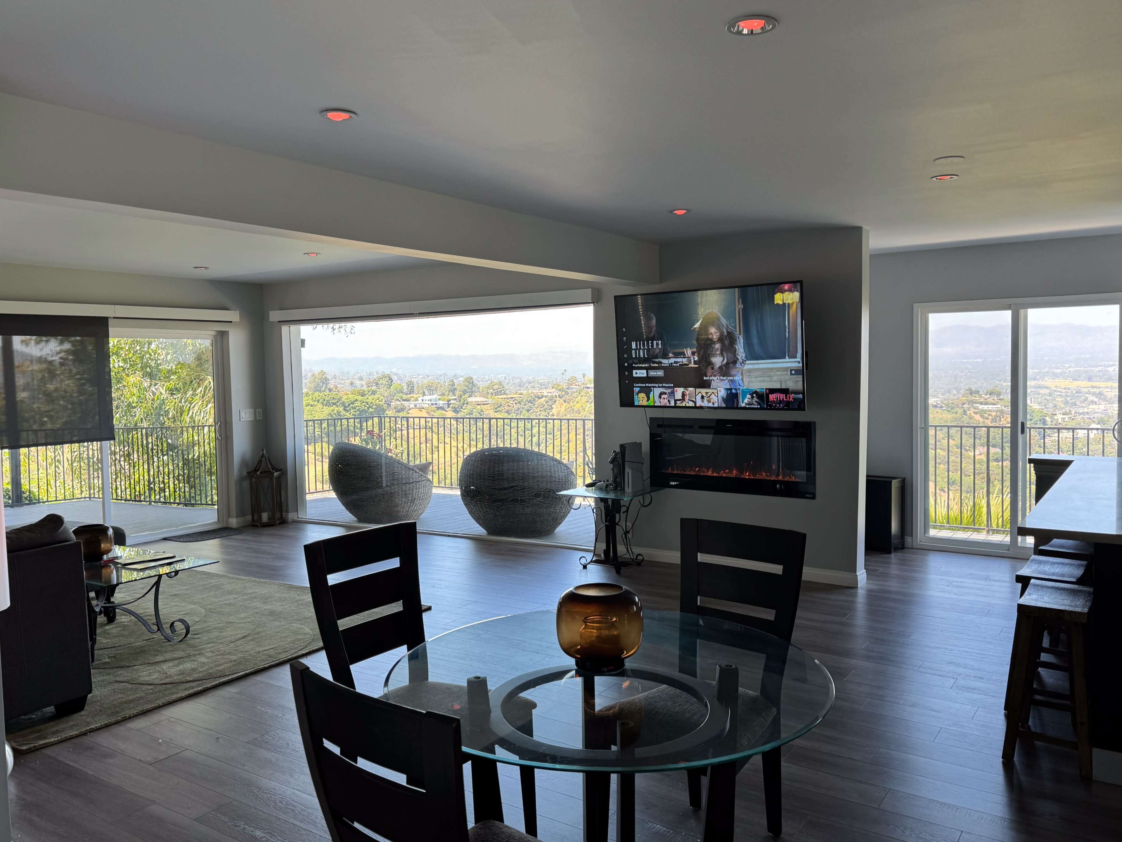 A modern living room with a glass table, black chairs, a wall-mounted TV, and a fireplace, featuring large windows that overlook a scenic view.