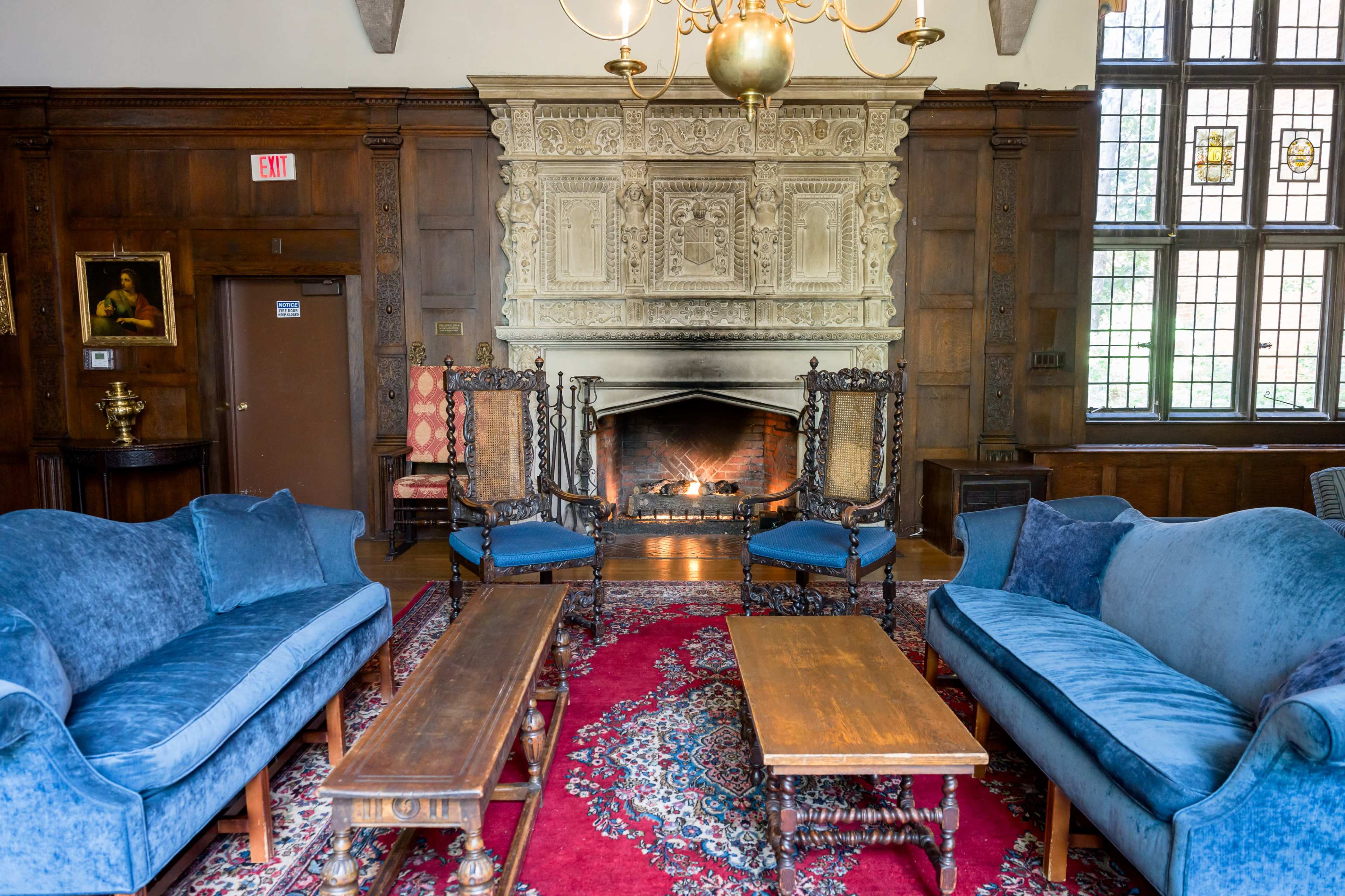 A cozy living room with blue sofas and wooden tables facing a fireplace, flanked by two ornate chairs in front of a detailed stone wall.