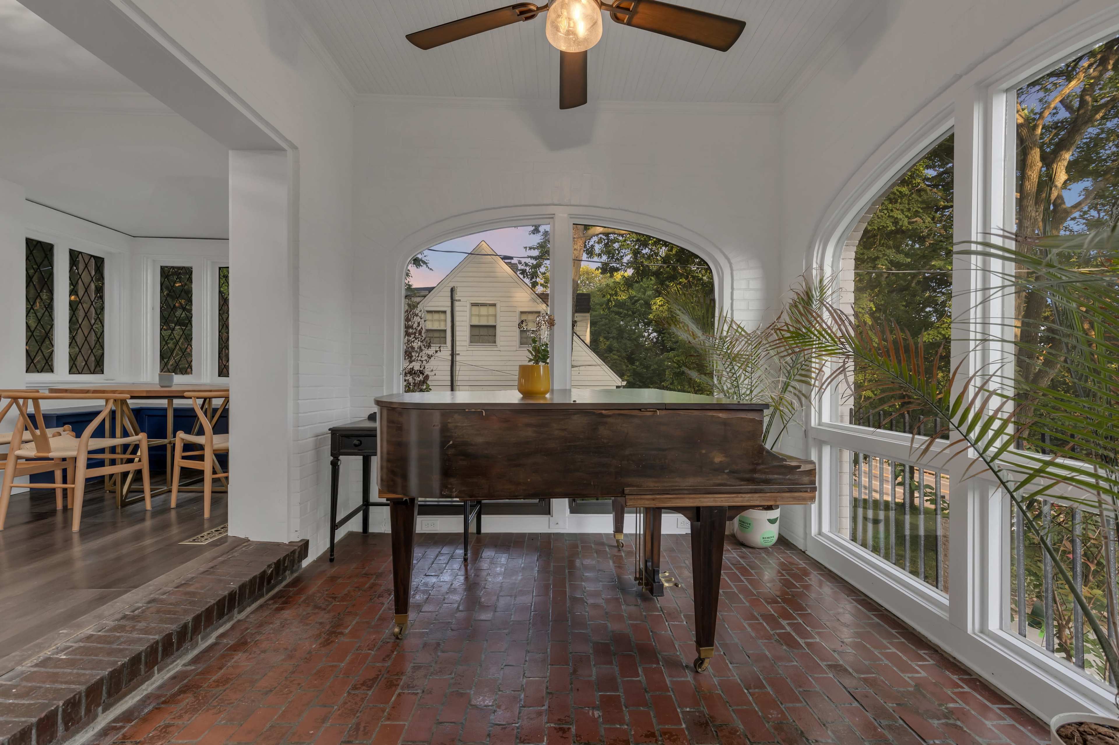 A grand piano is positioned in a sunlit room with arched windows and a view of a house surrounded by trees.