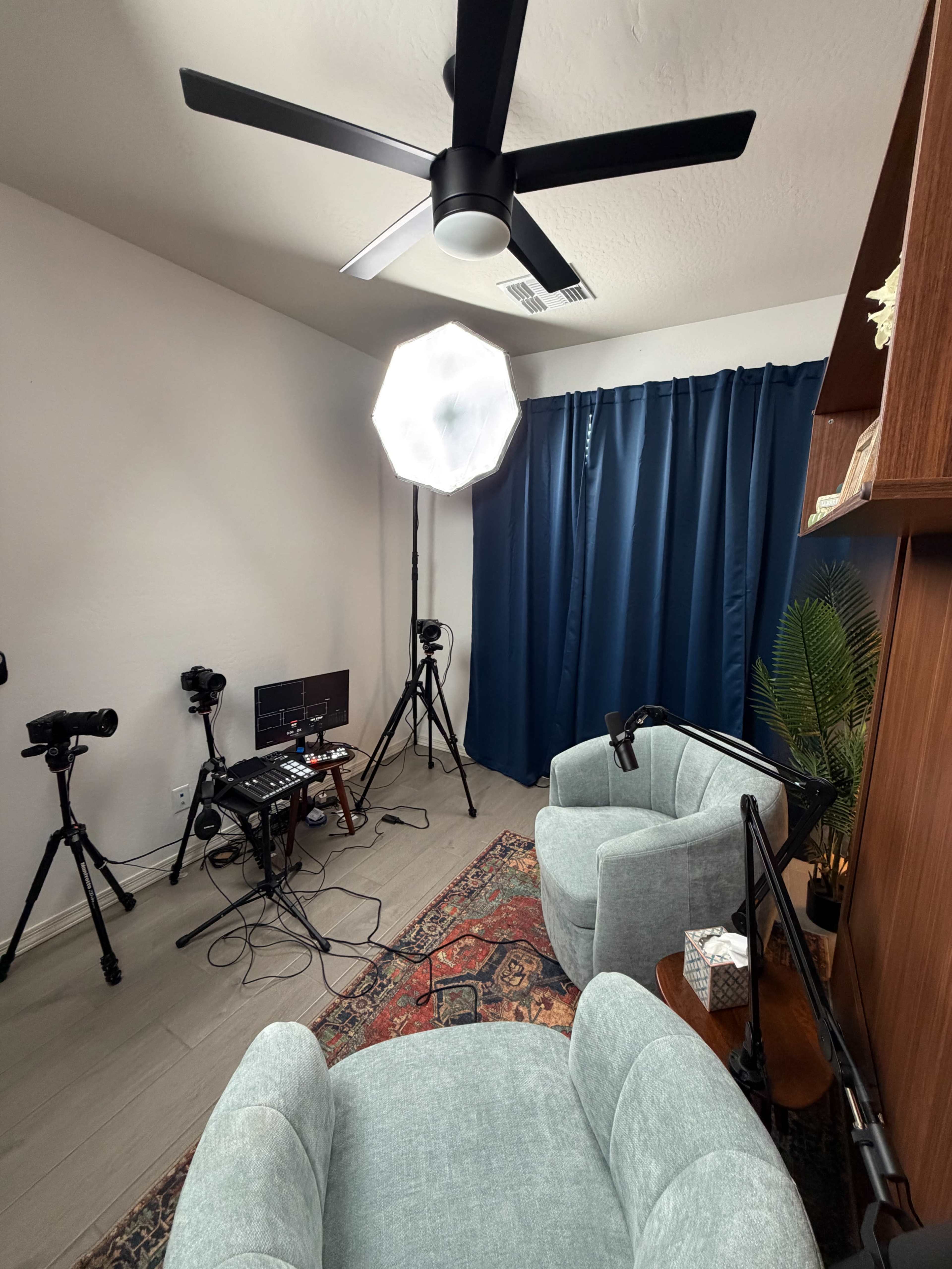 The image shows a small studio setup featuring two light blue chairs, a light umbrella, multiple cameras on tripods, and a small table with equipment, all arranged in a room with a dark blue curtain.