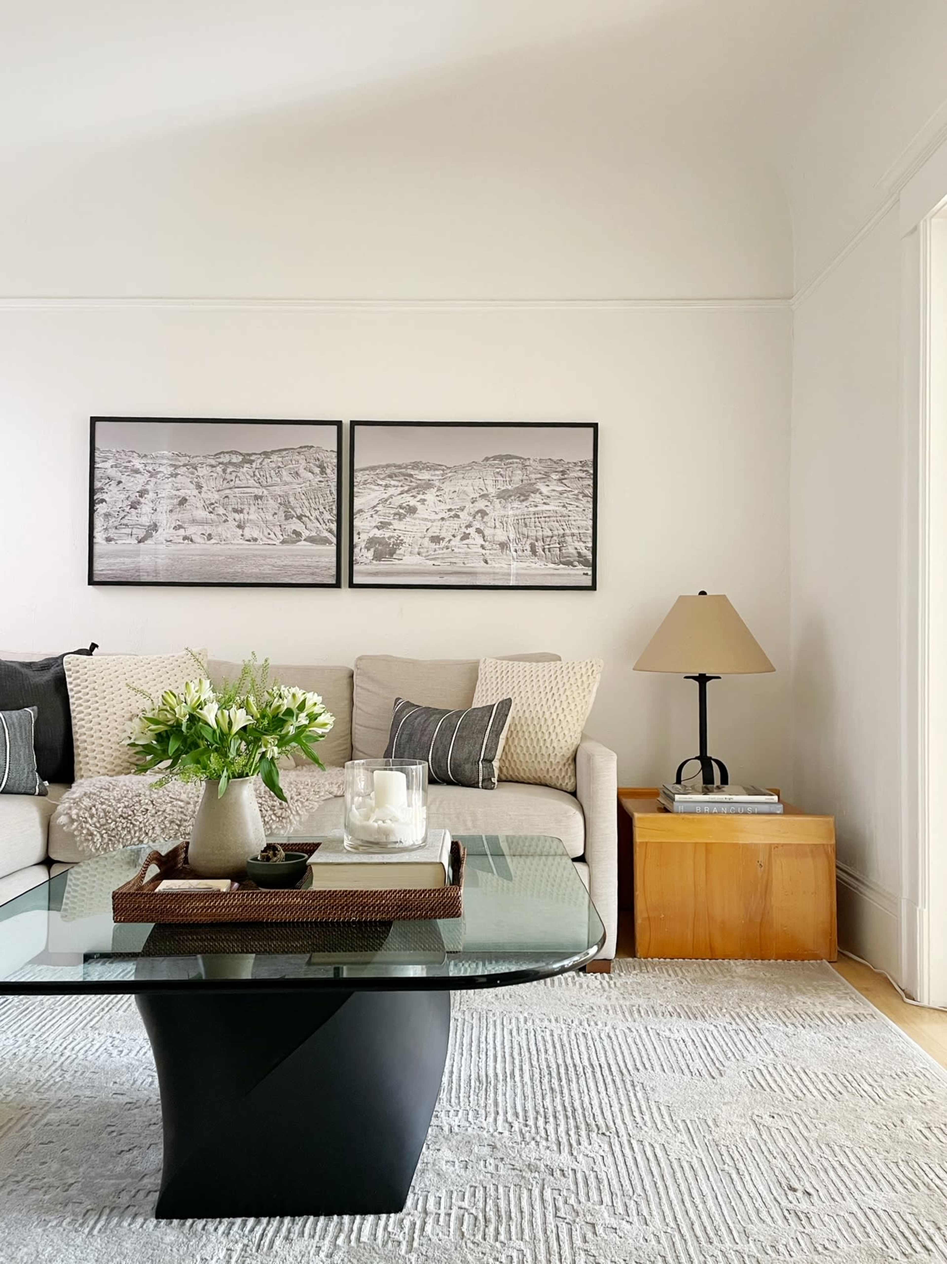 A modern living room with a glass coffee table, a beige sofa adorned with patterned cushions, two framed black-and-white photographs on the wall, and a wooden side table with a lamp.