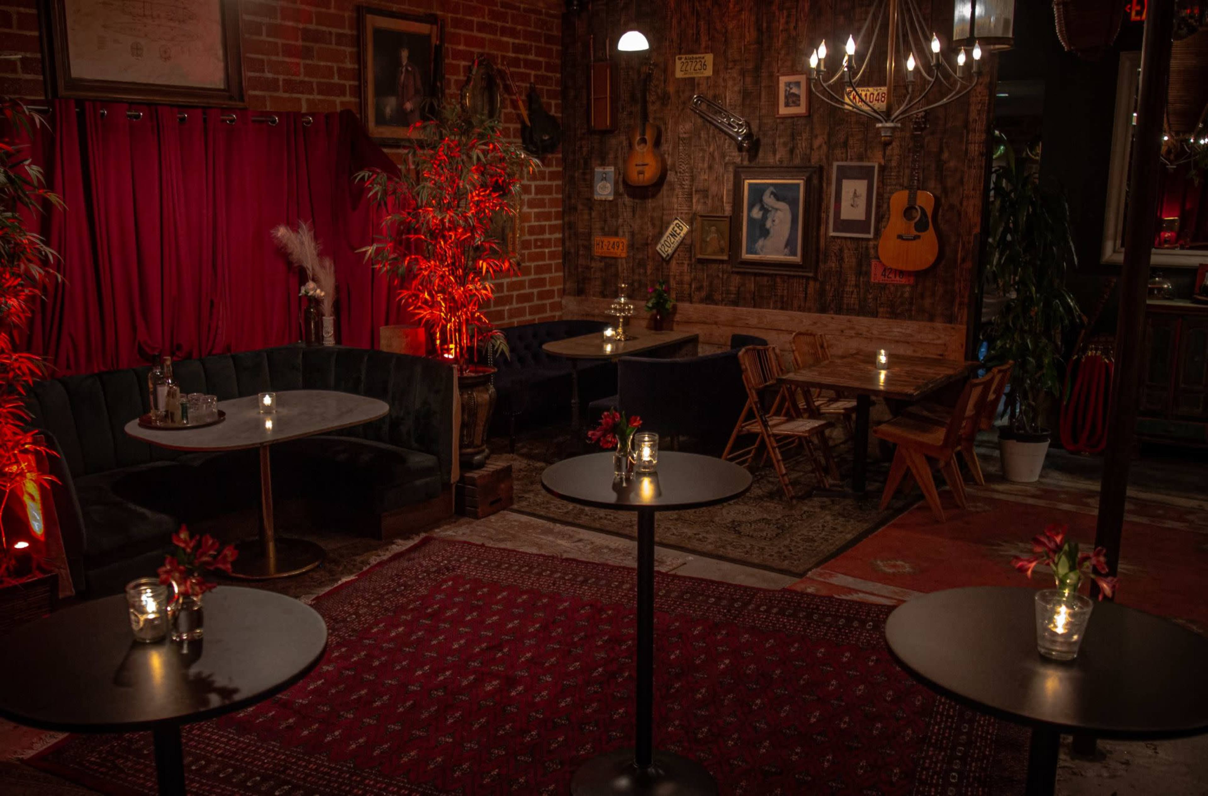 The image depicts a cozy indoor lounge area featuring dark green seating, wooden tables, and low lighting, accented by red plants and wall decorations.