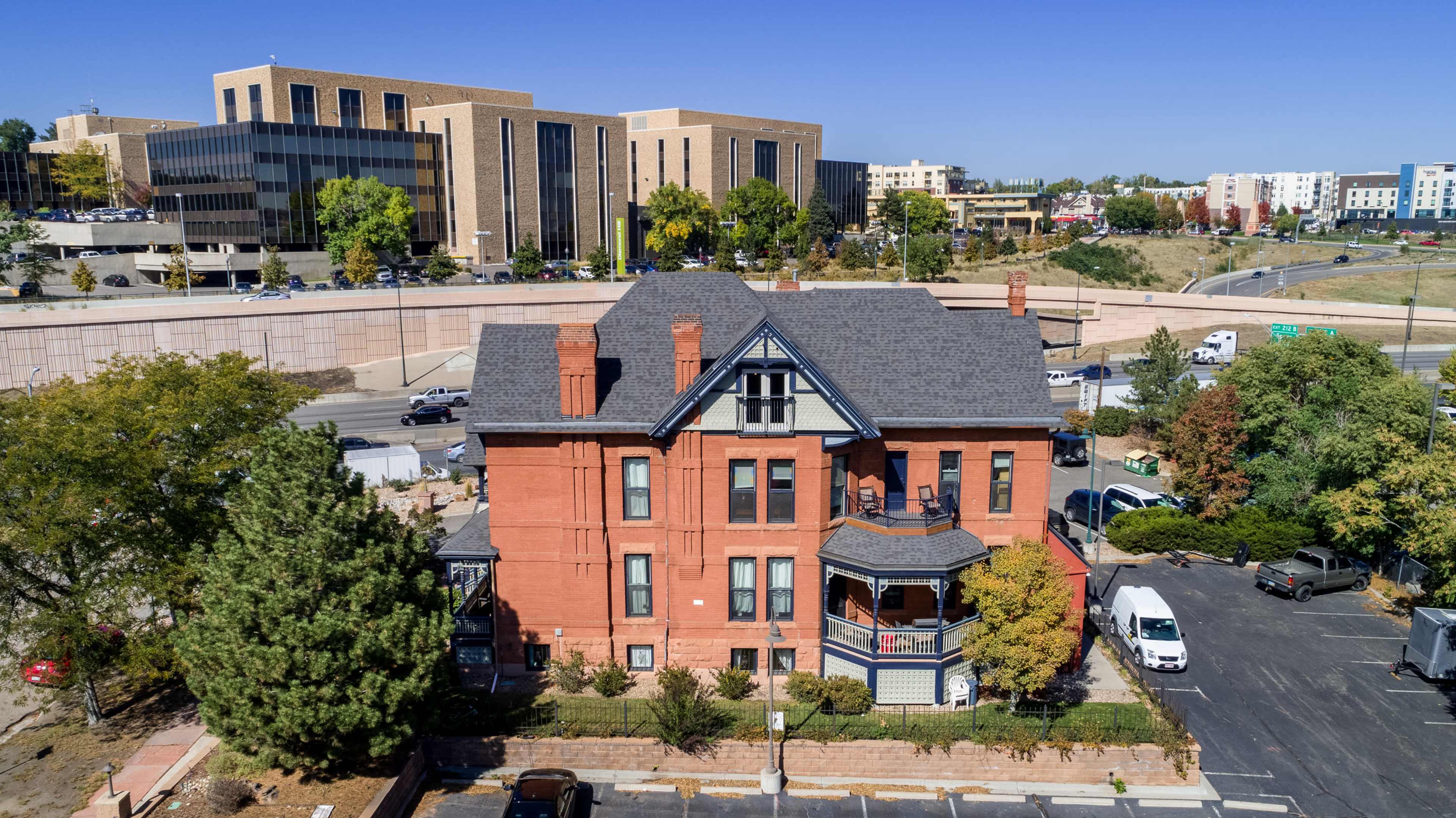 A large brick Victorian house stands in the foreground, surrounded by trees and parked cars, with modern buildings and a highway in the background.