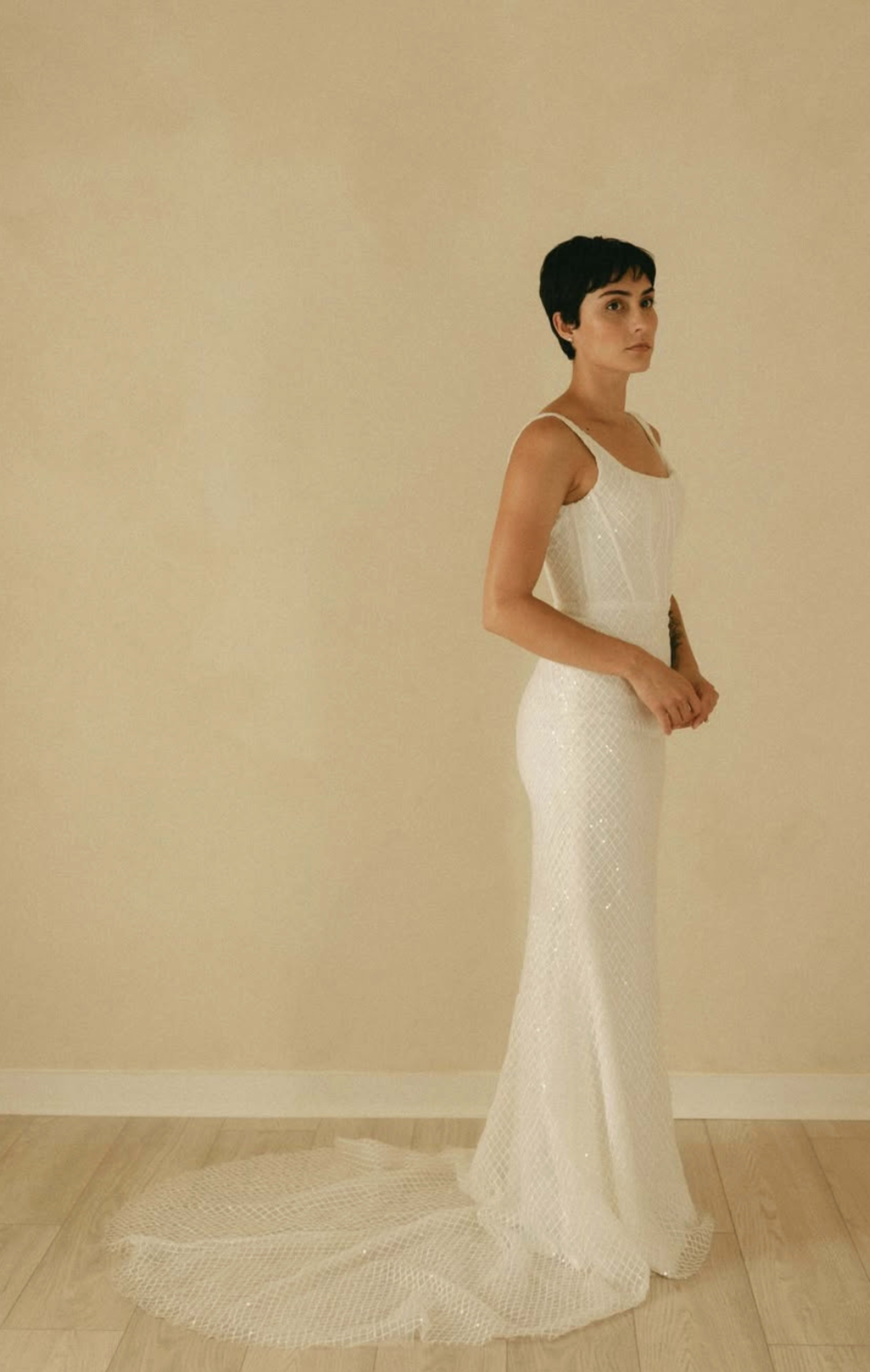 A woman stands in profile wearing a fitted white dress with a train against a neutral background.