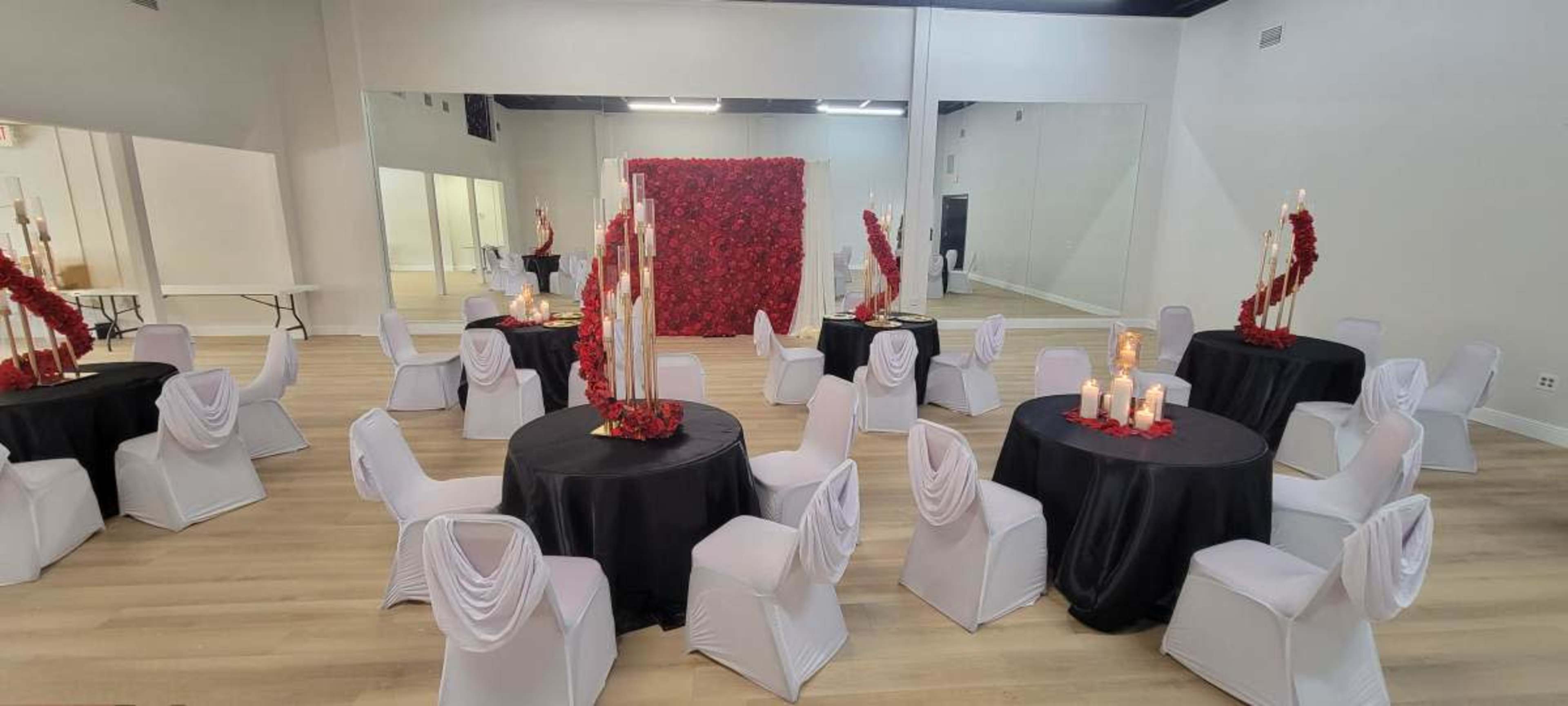 A spacious event venue arranged with round tables covered in black cloth, white chairs with draped backs, and a backdrop of red roses.