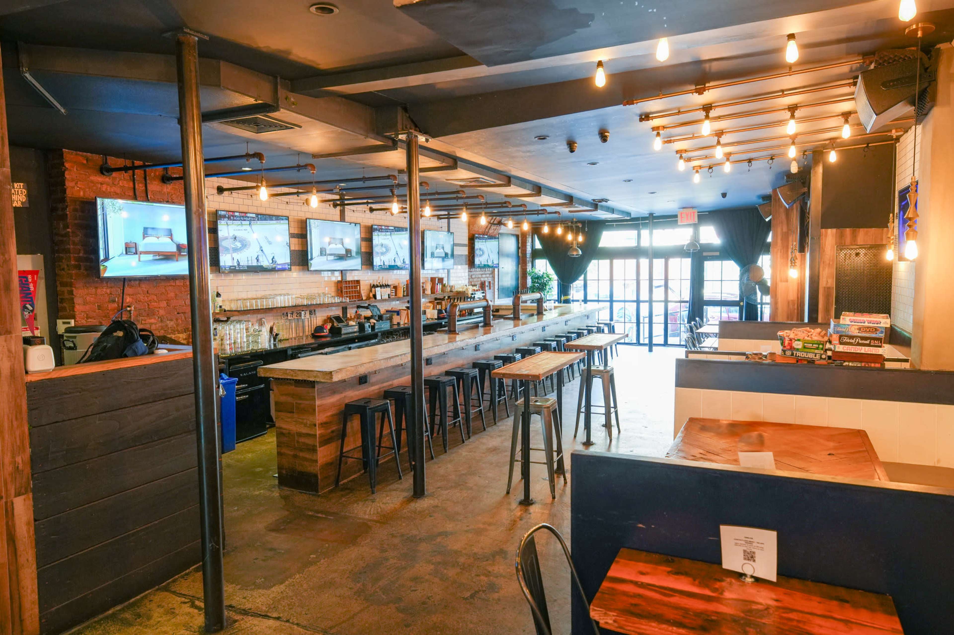 The image shows the interior of a bar or restaurant with wooden countertops, high stools, multiple televisions on the wall, and a spacious layout that leads to a door with natural light.
