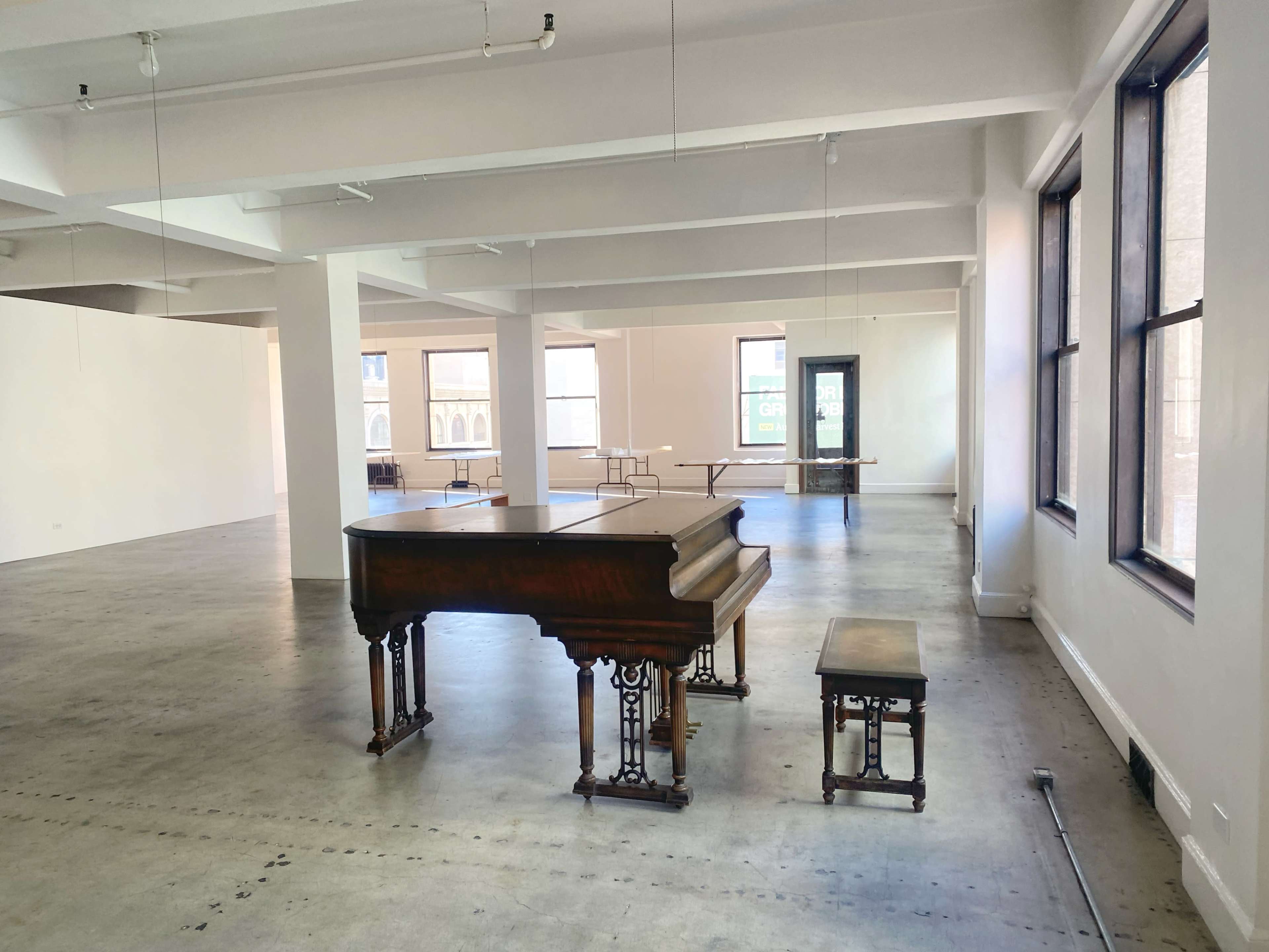 The image shows an empty, spacious room with a grand piano and a matching bench placed near large windows.