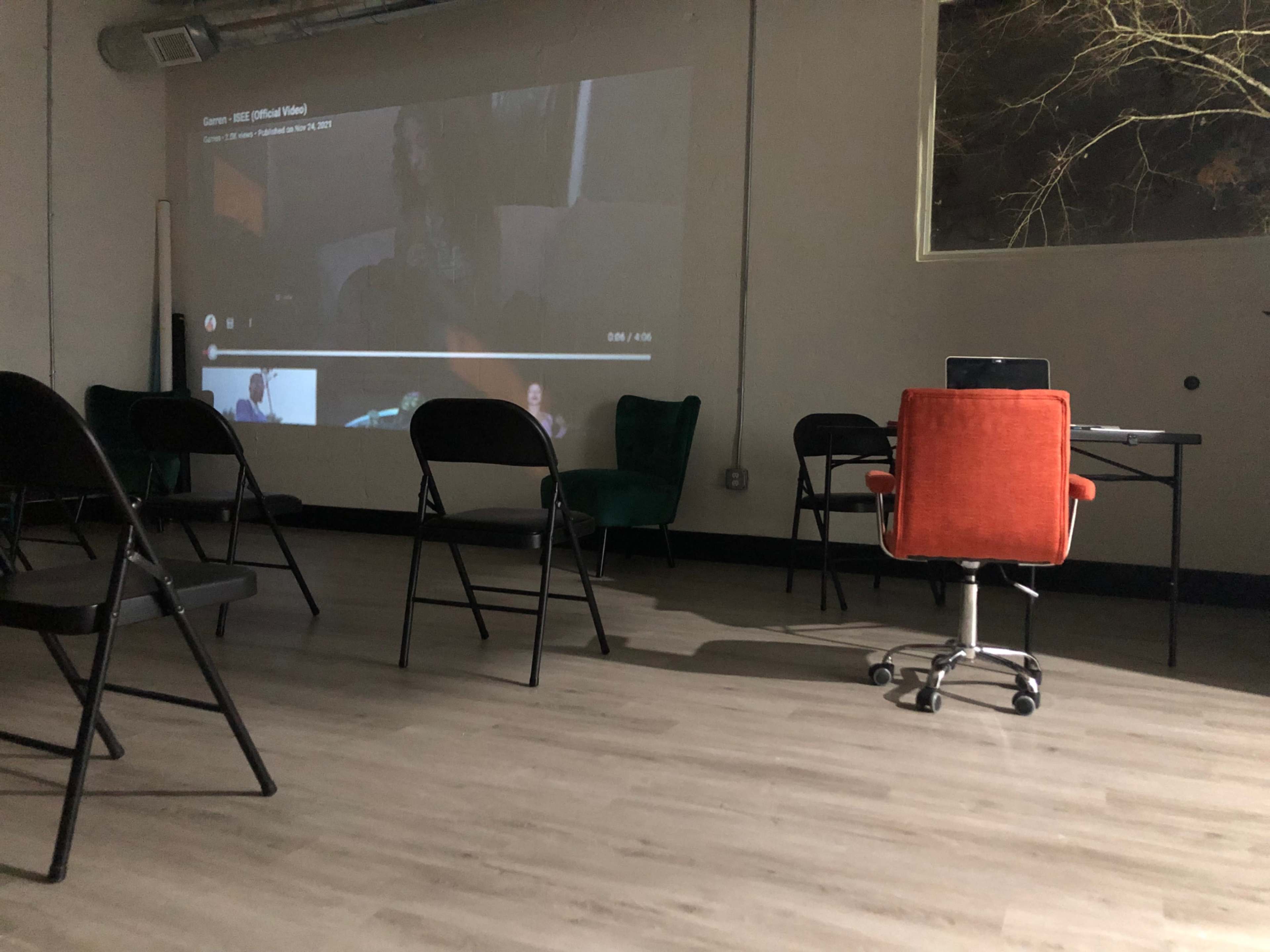A video is projected onto a wall in a sparsely furnished room with several empty chairs and a desk.