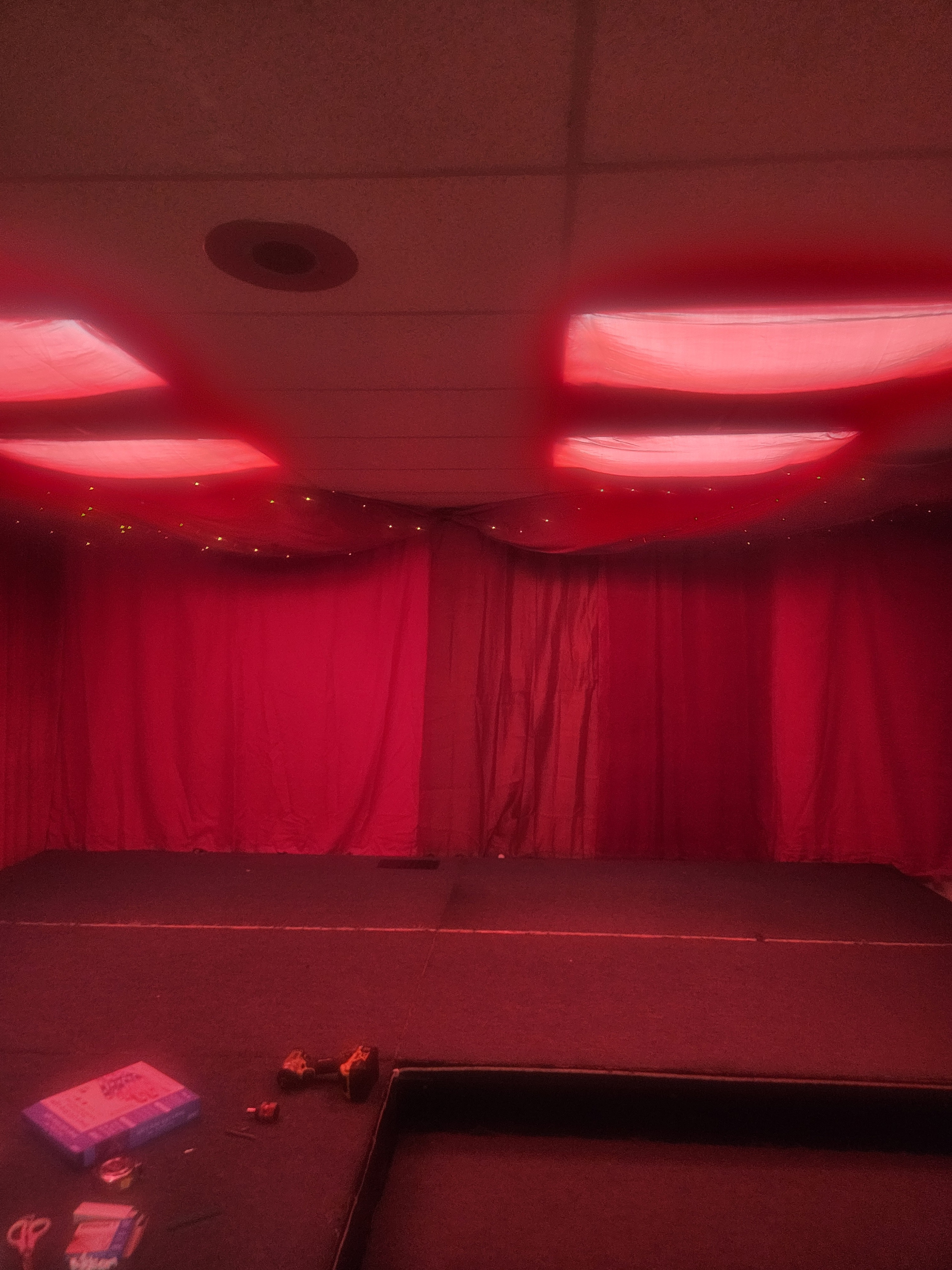 The image shows a stage area with red draped curtains and dim lighting, featuring a dark floor and scattered tools in the foreground.