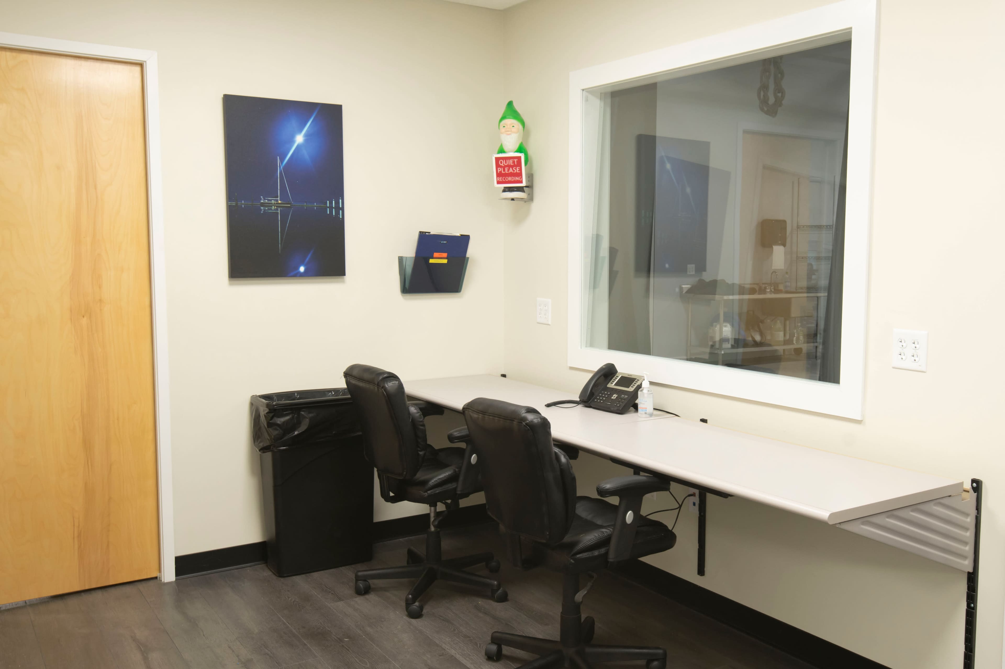 A small office space with two black chairs positioned at a gray desk, a telephone, a trash can, and a window with a framed picture on the wall.
