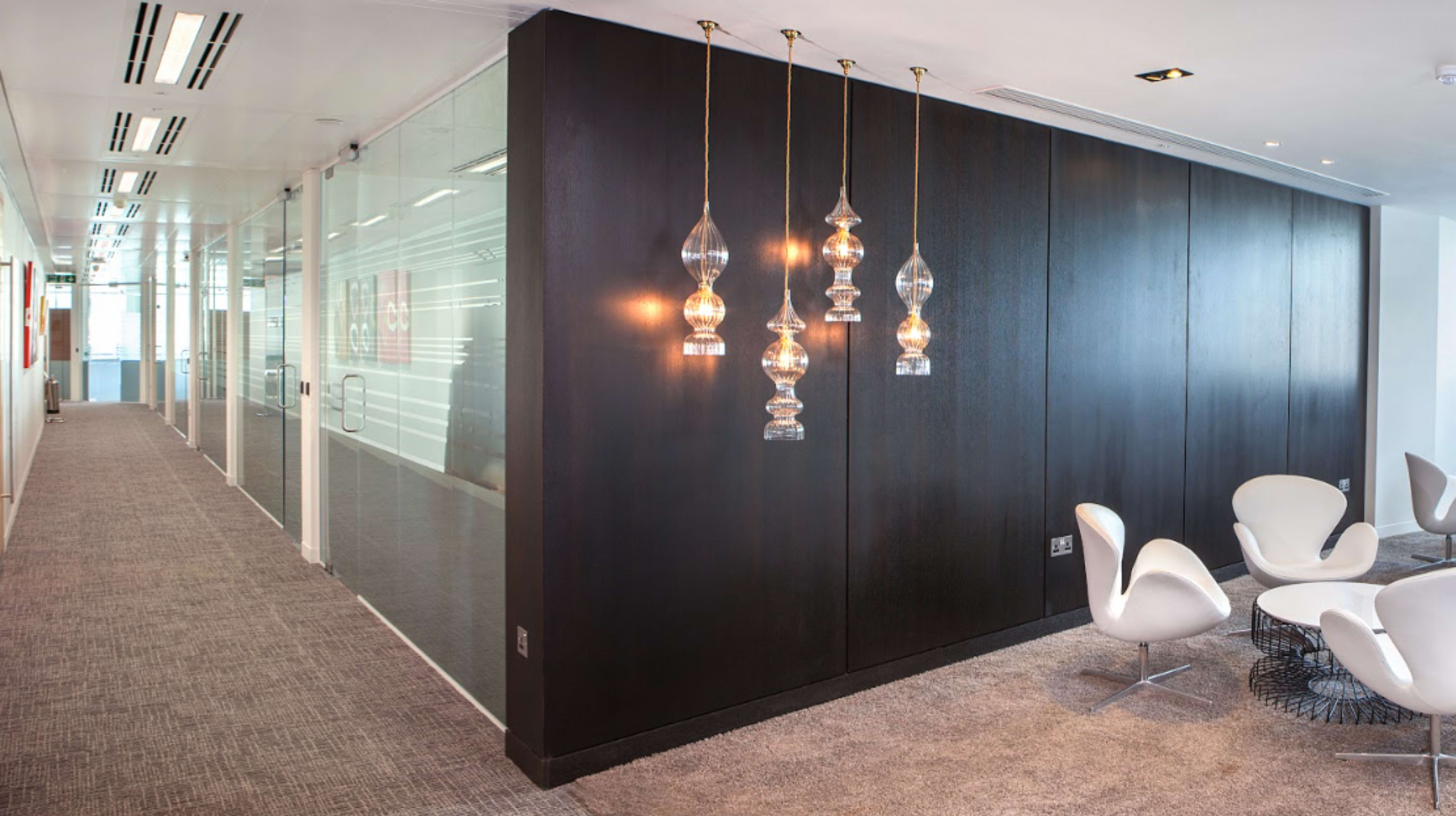 A modern office interior with a dark wall, glass partitions, and a seating area featuring white chairs and hanging light fixtures.