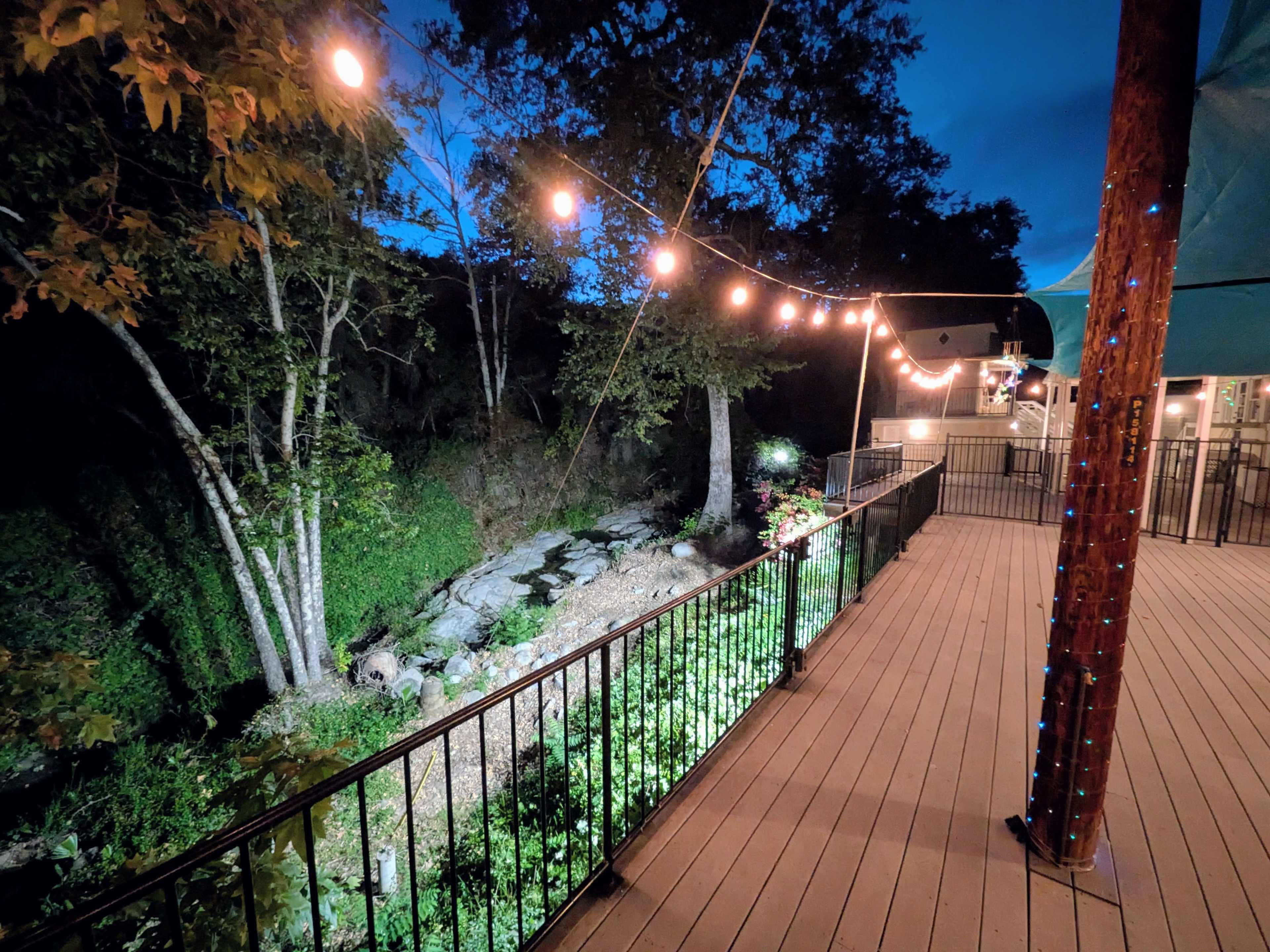 A deck with string lights overlooks a creek bordered by trees, set against a twilight sky.