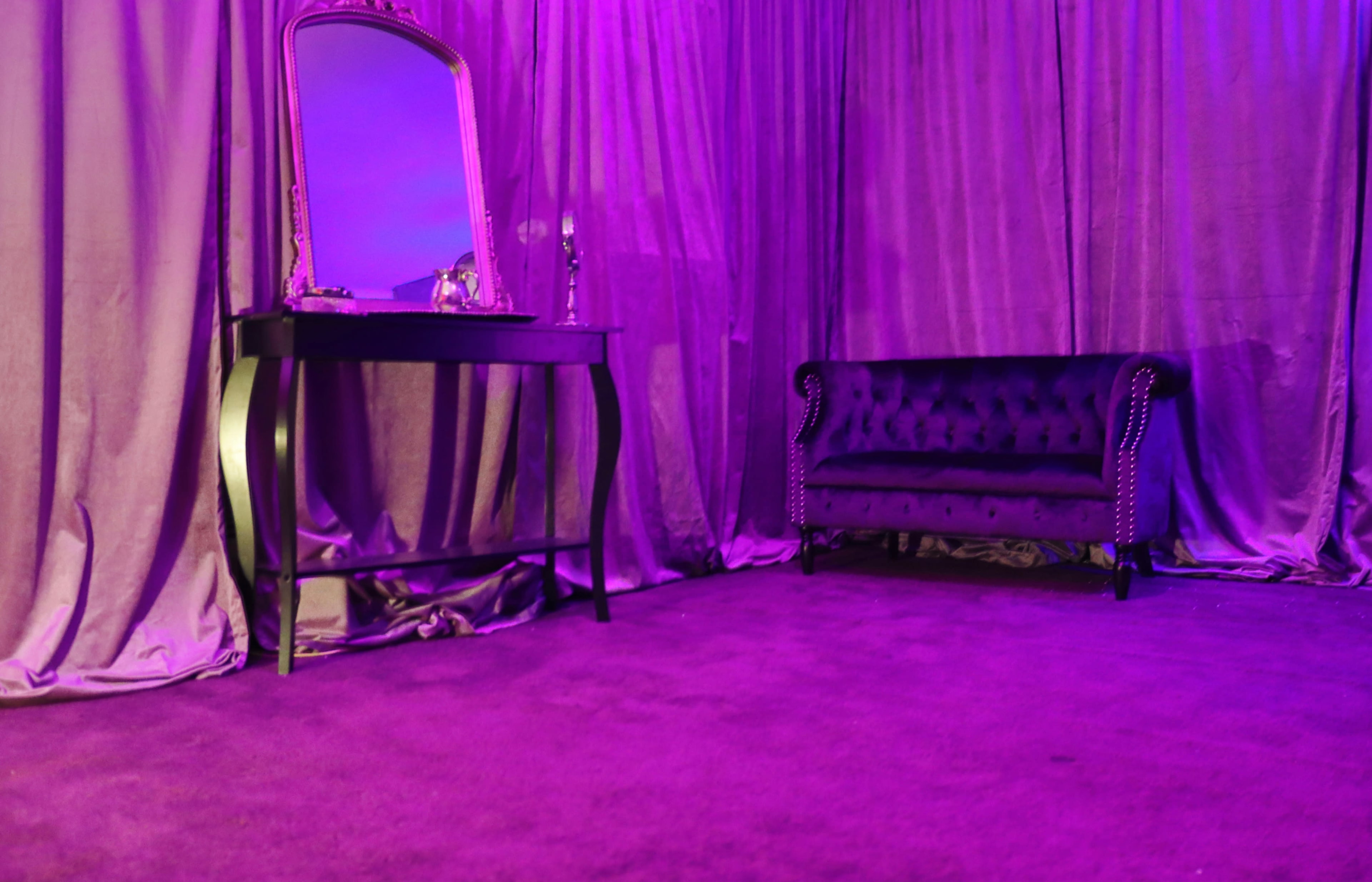 A dark purple room features a velvet sofa and a mirrored vanity in front of draped curtains.