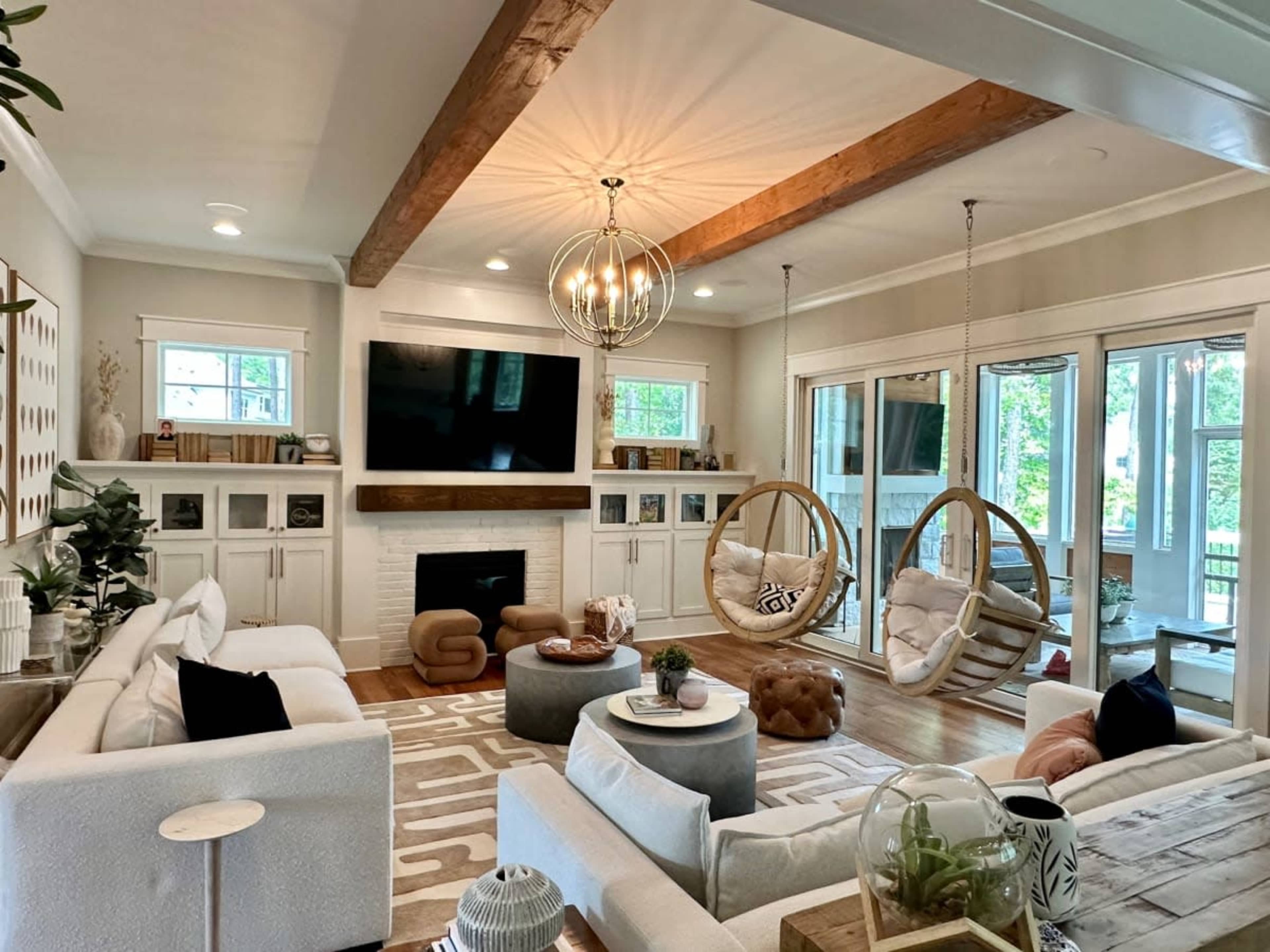 A modern living room features a large TV mounted above a white fireplace, accompanied by two hanging chairs and plenty of natural light from the surrounding windows.