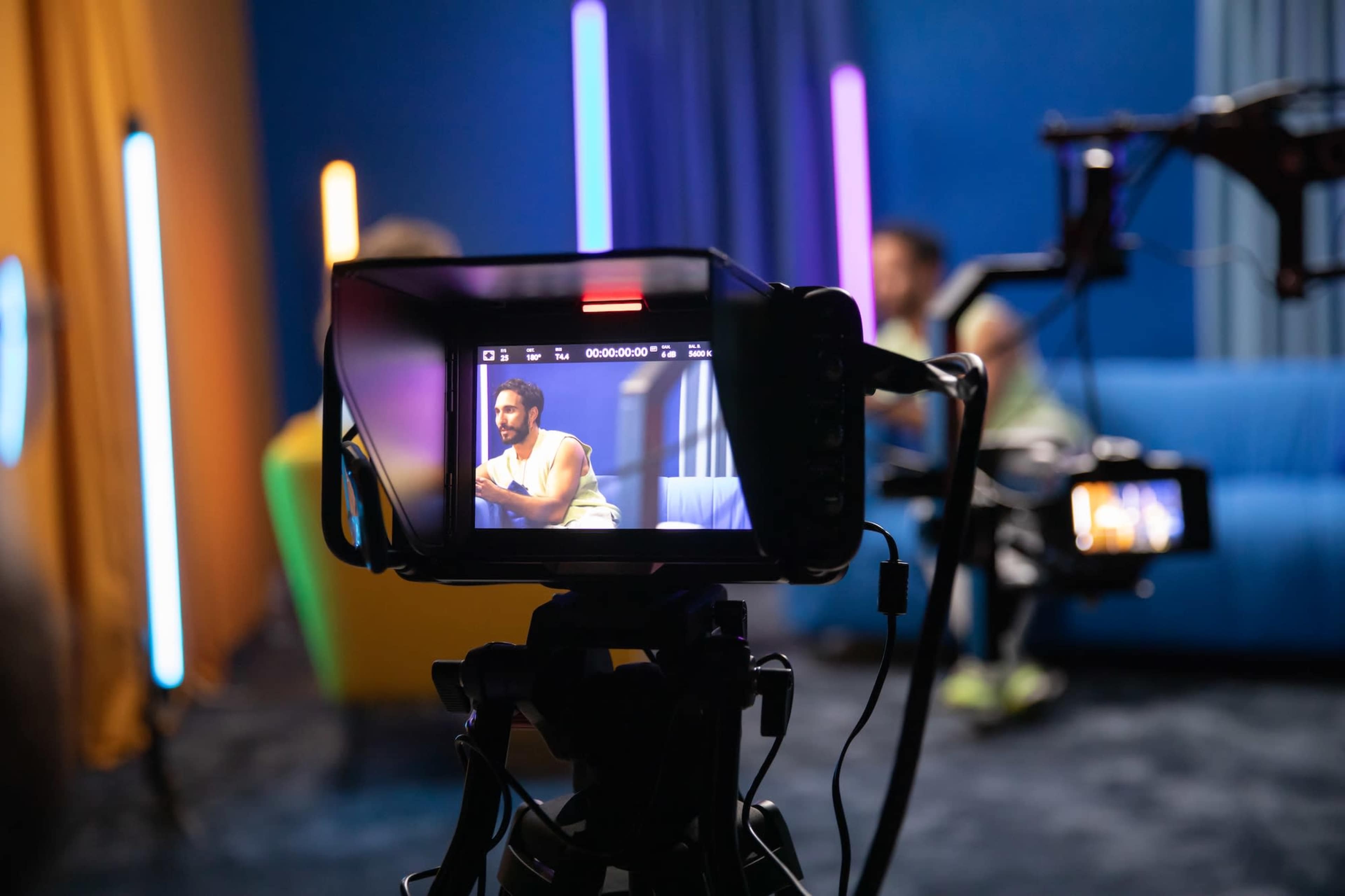 A camera captures a man speaking while seated on a colorful couch in a studio with bright lighting.