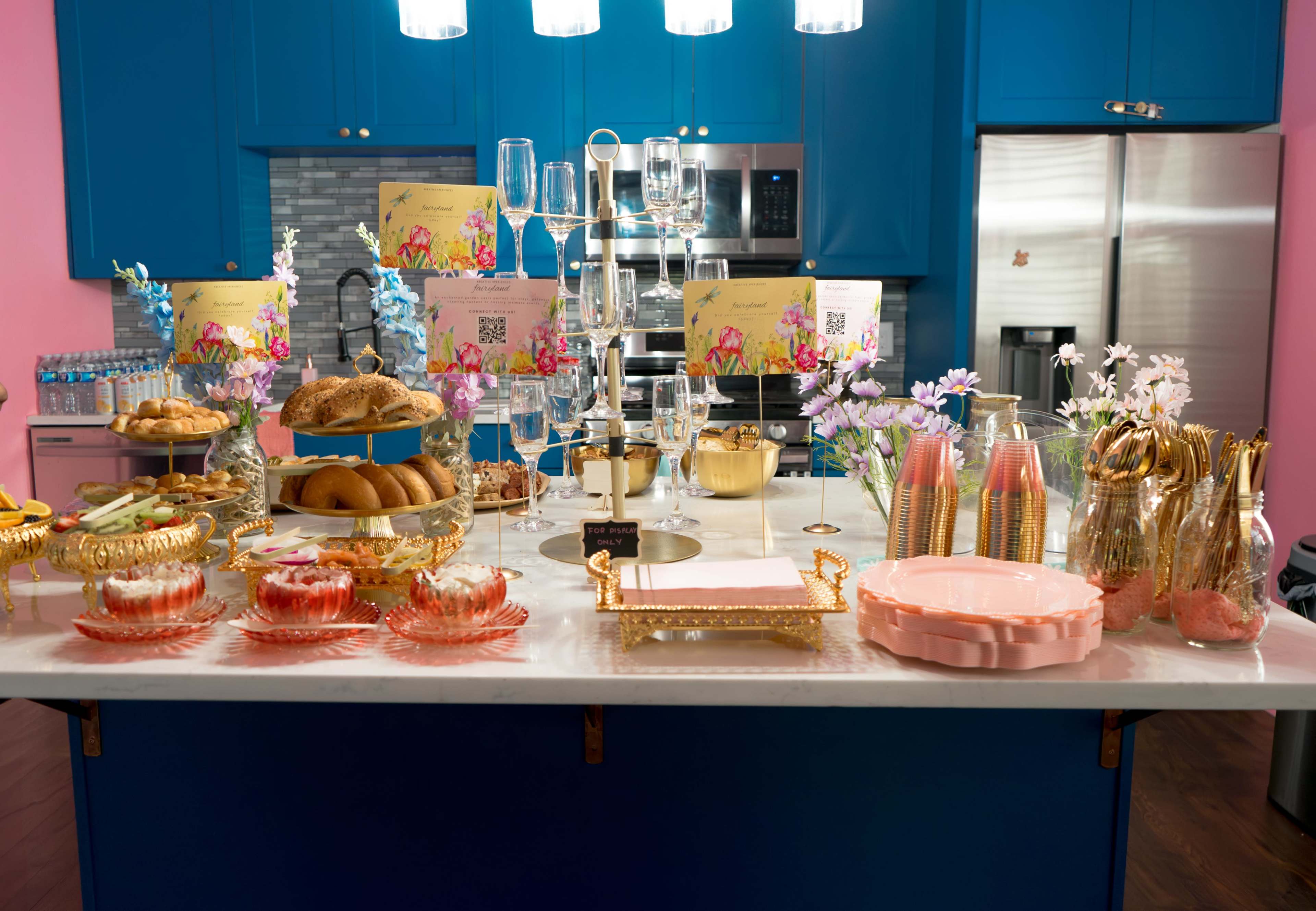 The image shows a colorful kitchen island adorned with various baked goods, drinks, and decorative tableware, set against bright blue cabinets and pink walls.