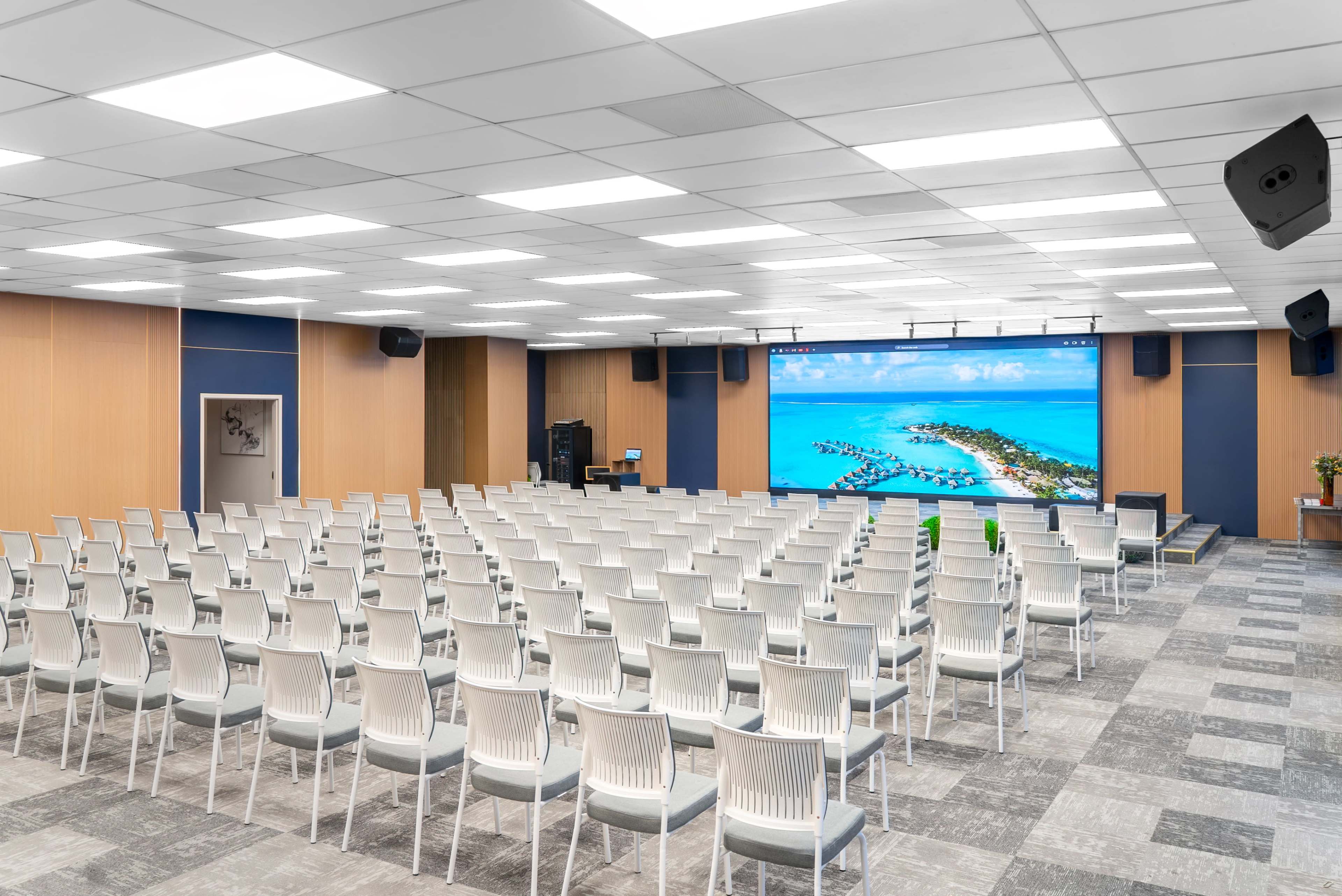 The image shows a large conference room filled with rows of white chairs facing a large screen displaying a tropical island scene.
