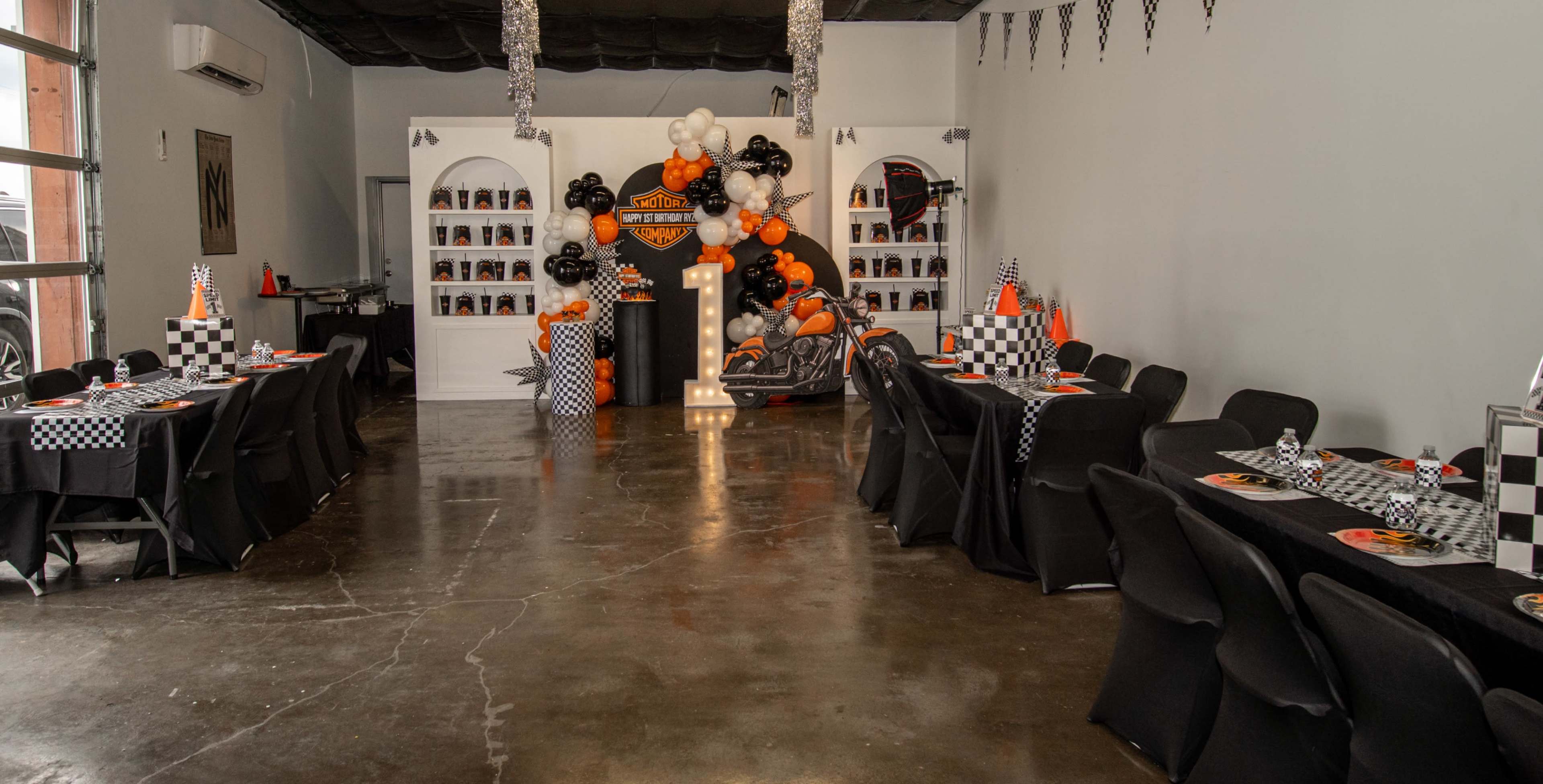 The image shows a party setup with black and orange decorations, featuring tables set up with checkered tablecloths and a large decorative display in the background that includes a large "1" sign and balloons.