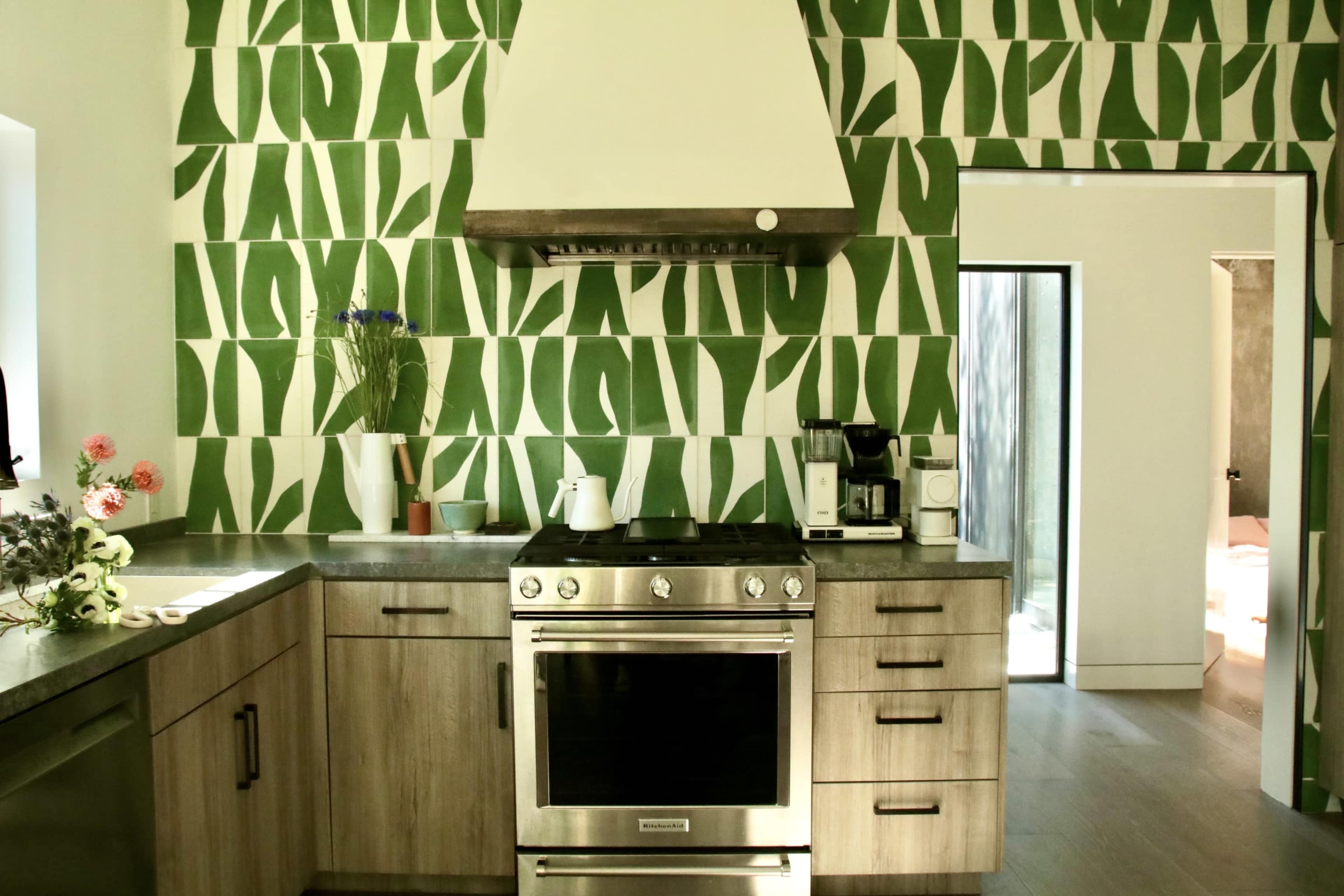 The kitchen features a stove beneath a hood, with a patterned green tile backsplash and wooden cabinets, and a doorway leading to another room in the background.