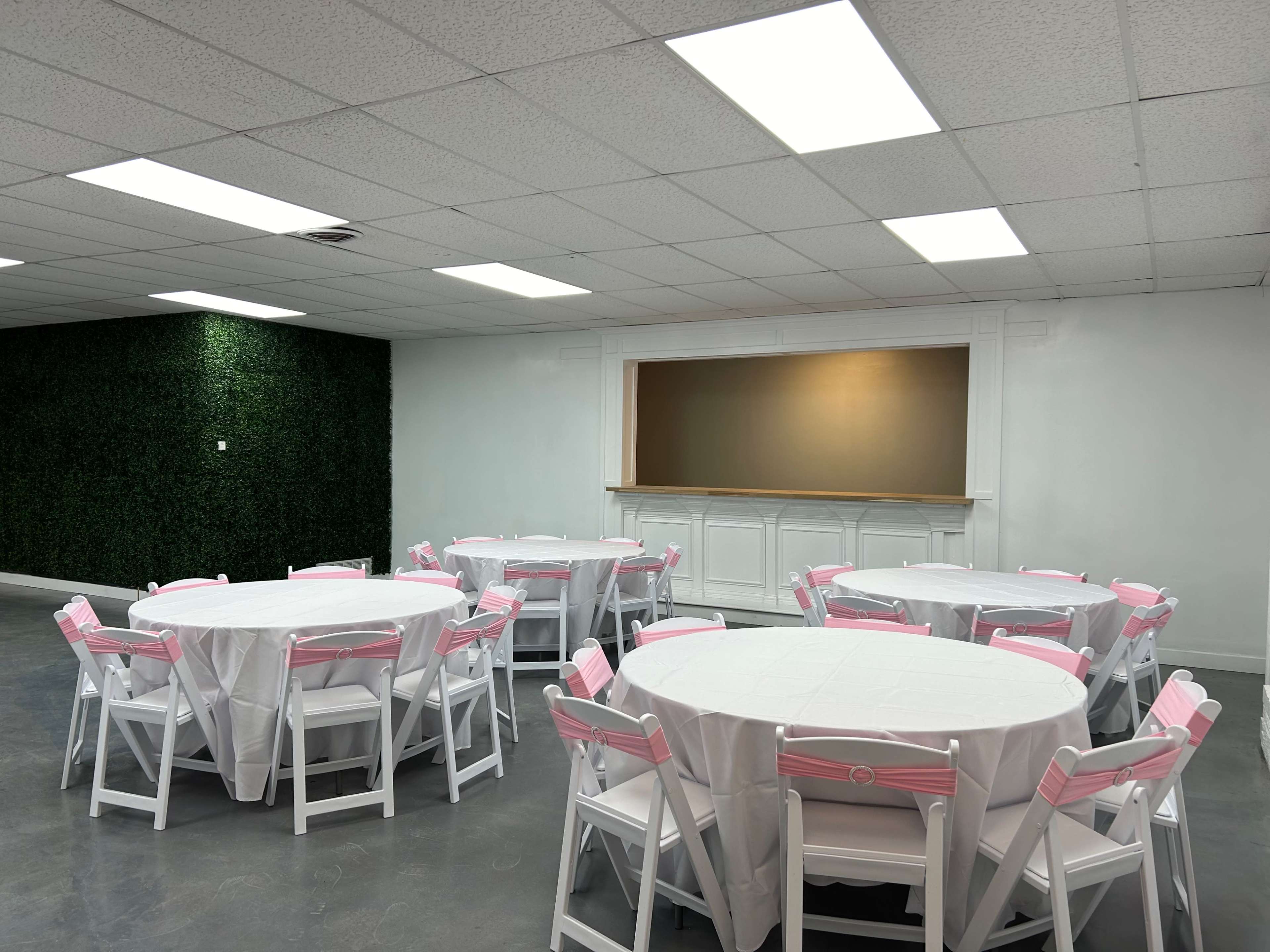 A spacious event room features round tables with white tablecloths and pink accents, arranged around a central area with a greenery wall and a sectioned off space at the front.