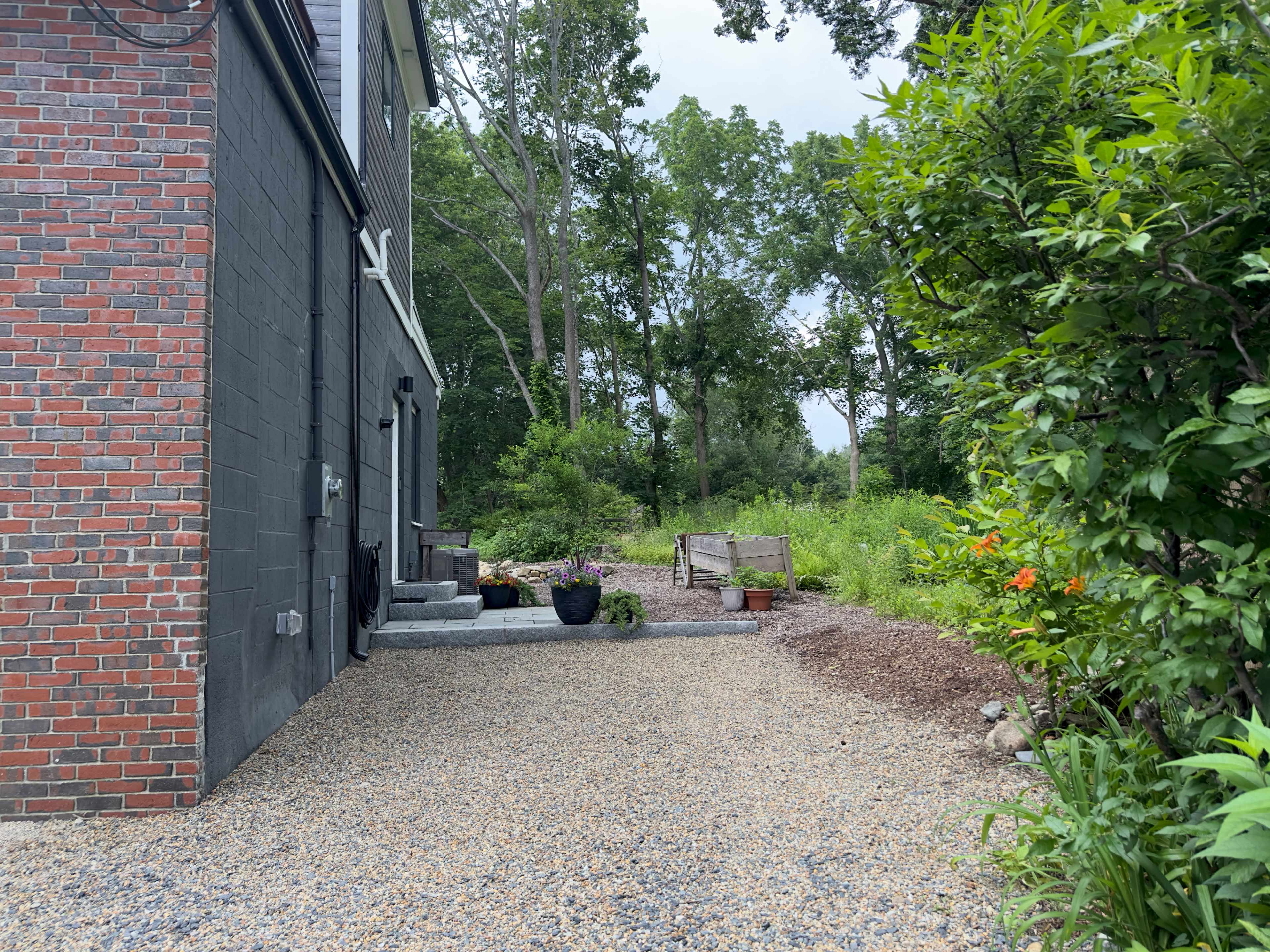 The scene shows a gravel path leading to a backyard area with plants and a wooden bench beside a house with a brick and black exterior.