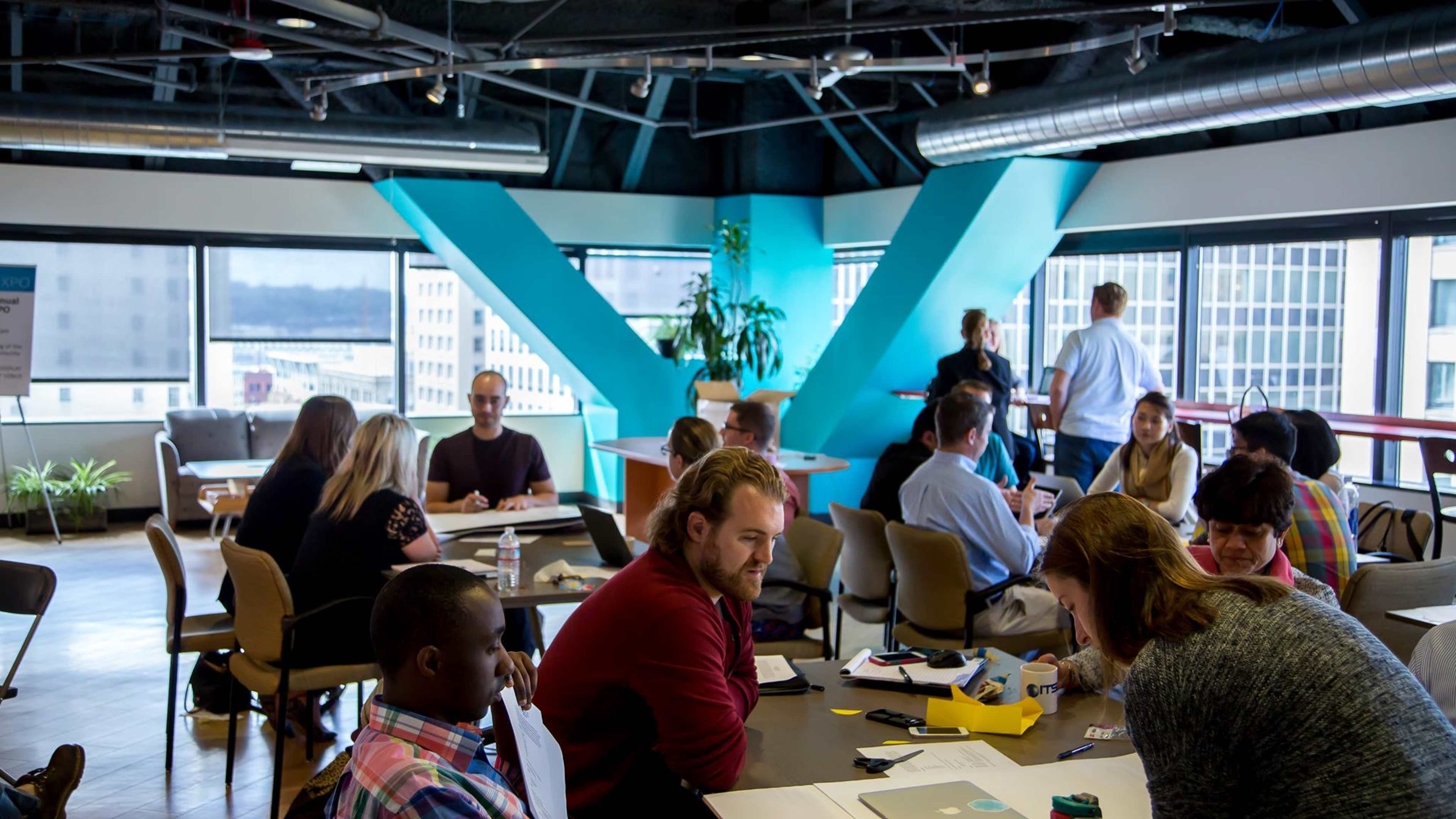 A group of people participates in a collaborative meeting in a modern office space with large windows and colorful decor.