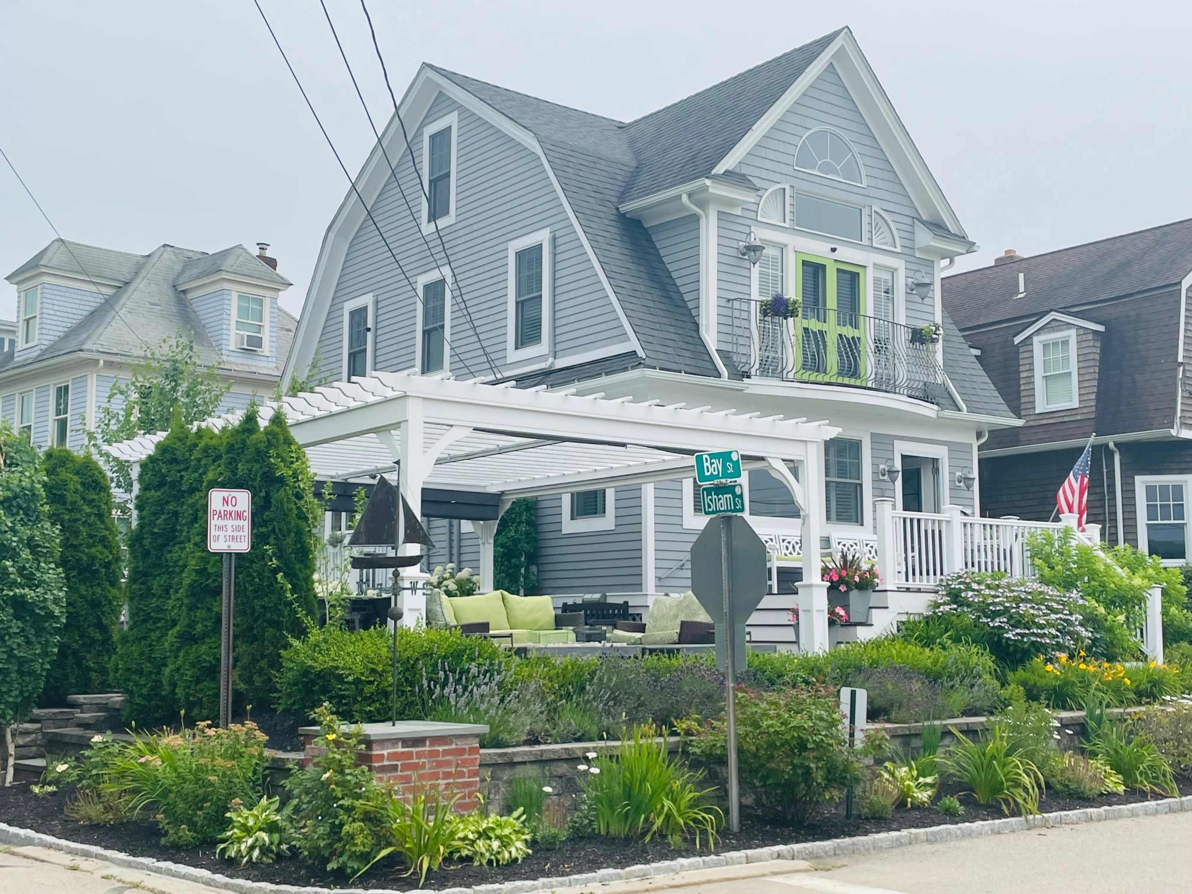 A gray two-story house with green accents features a pergola in the front yard, surrounded by landscaped gardens and a decorative brick wall.