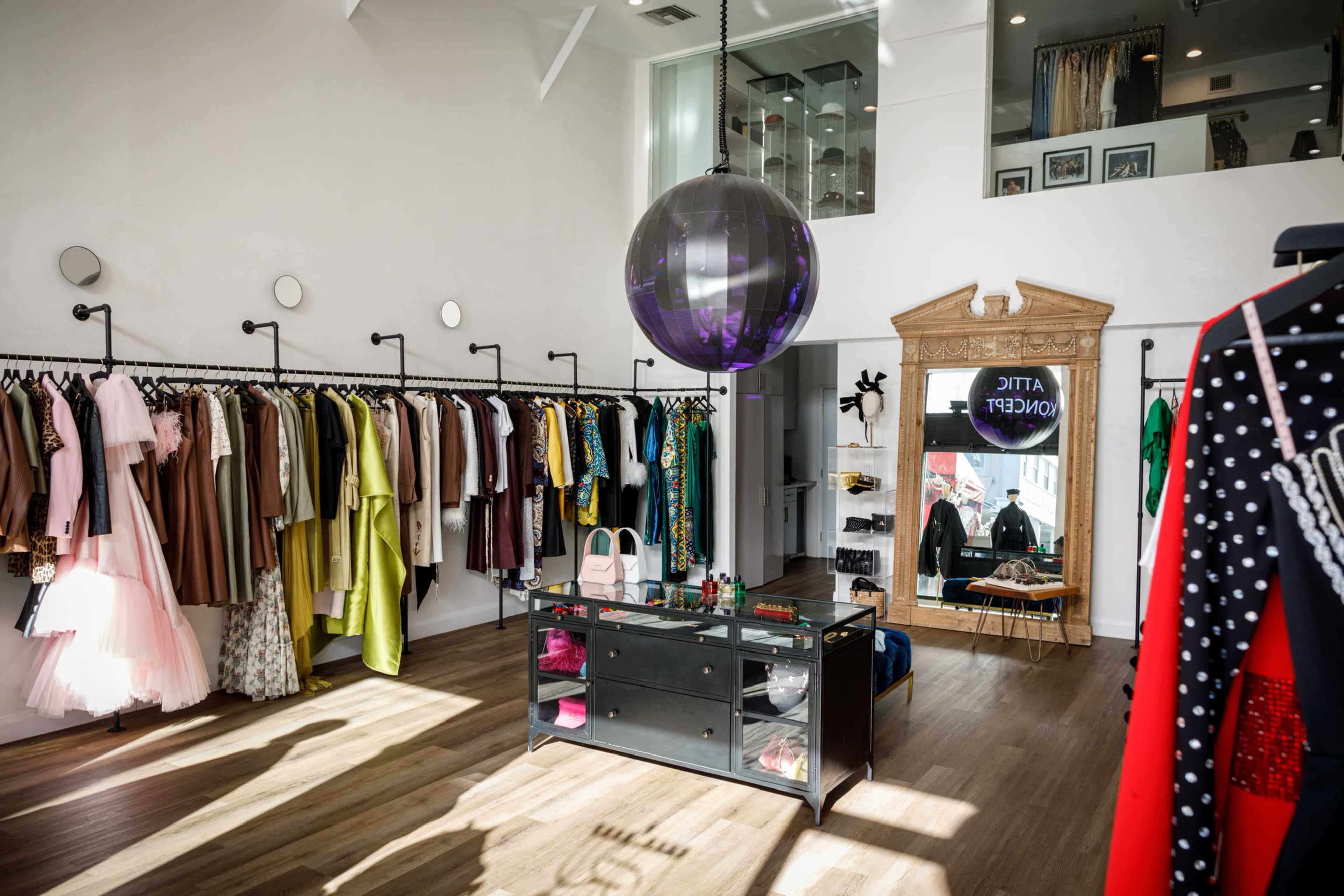 The image shows a stylish clothing boutique with racks of colorful garments lining the walls and a central display table featuring accessories.