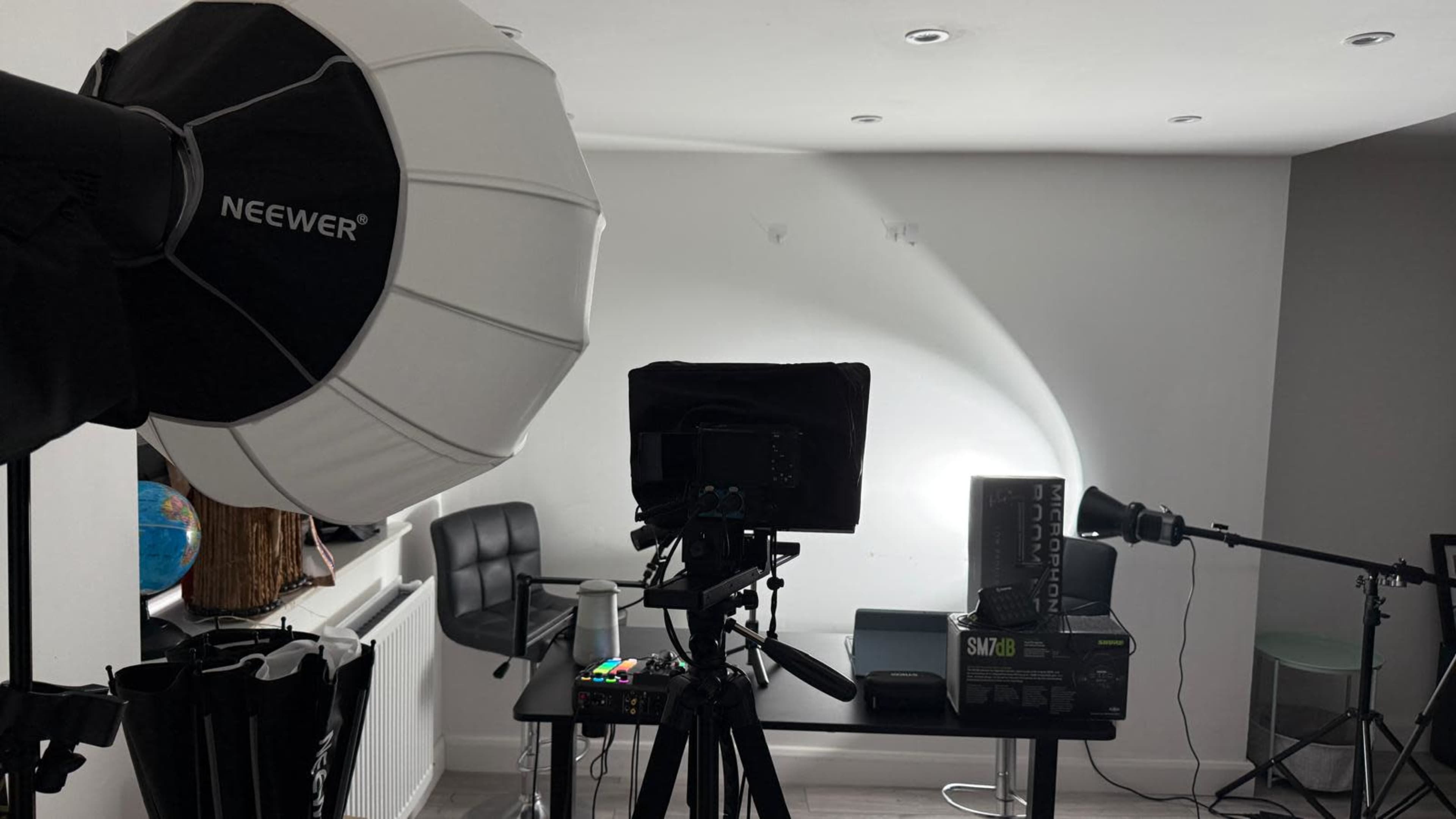 A recording studio setup features softbox lighting, a camera on a tripod, and a dark backdrop against a light wall.