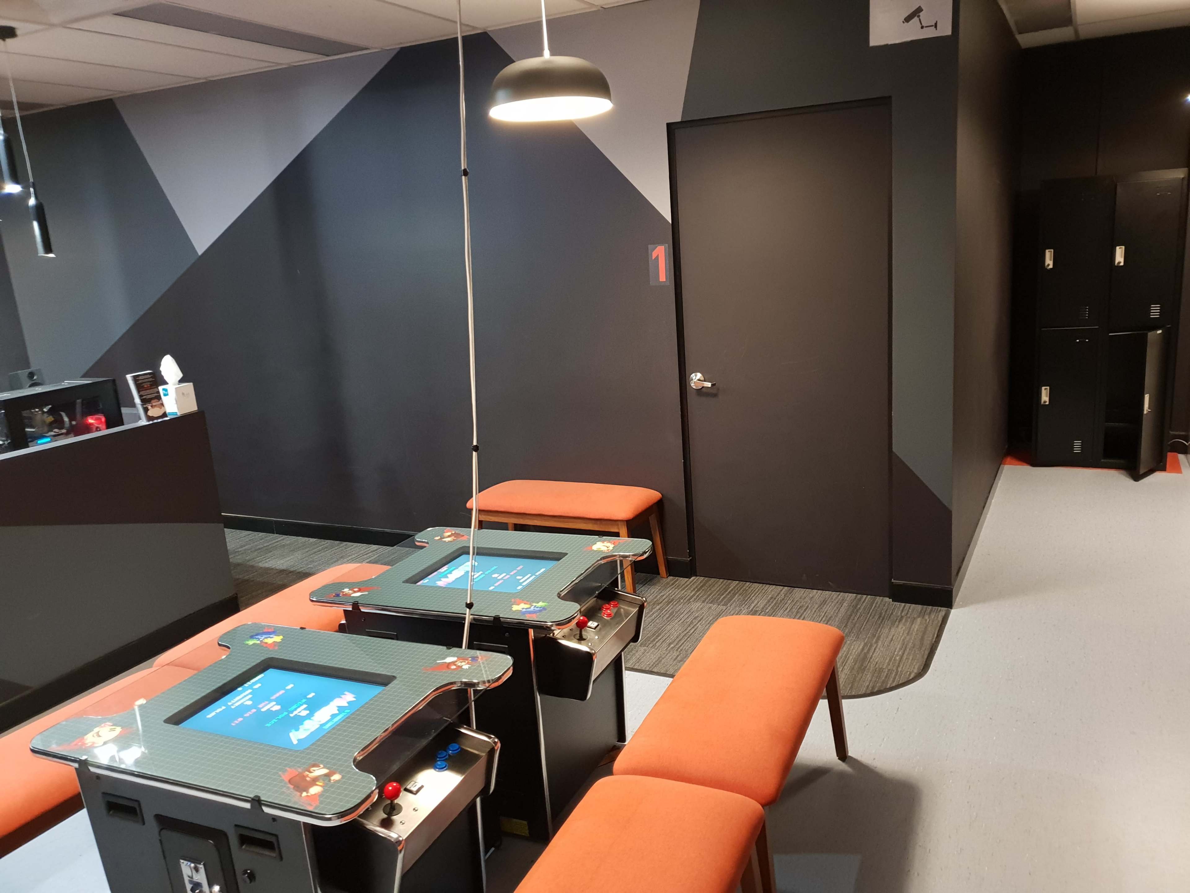 A gaming area with two arcade tables, orange benches, and a closed door in the background.