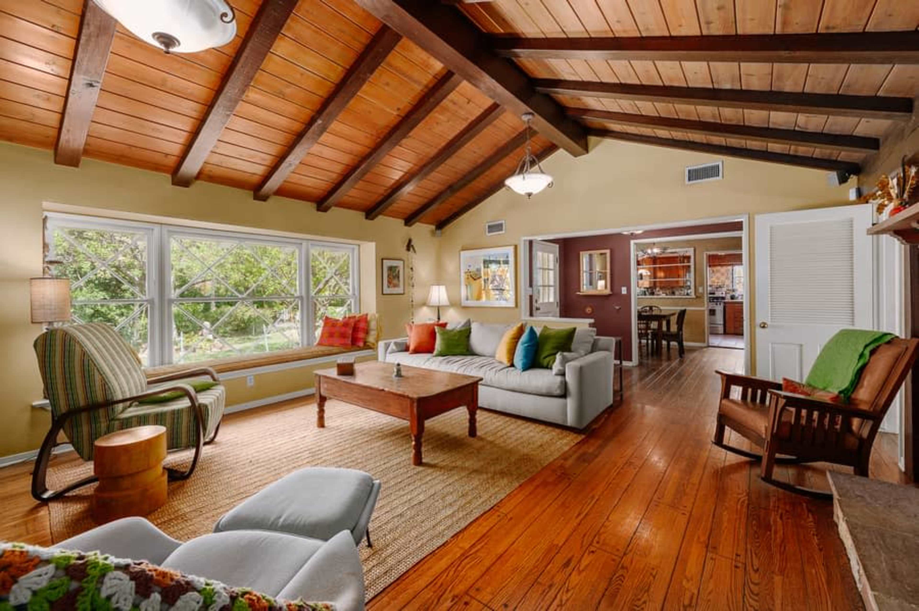The image shows a cozy living room with wooden beams, a sofa, multiple armchairs, and a view of a garden through large windows.
