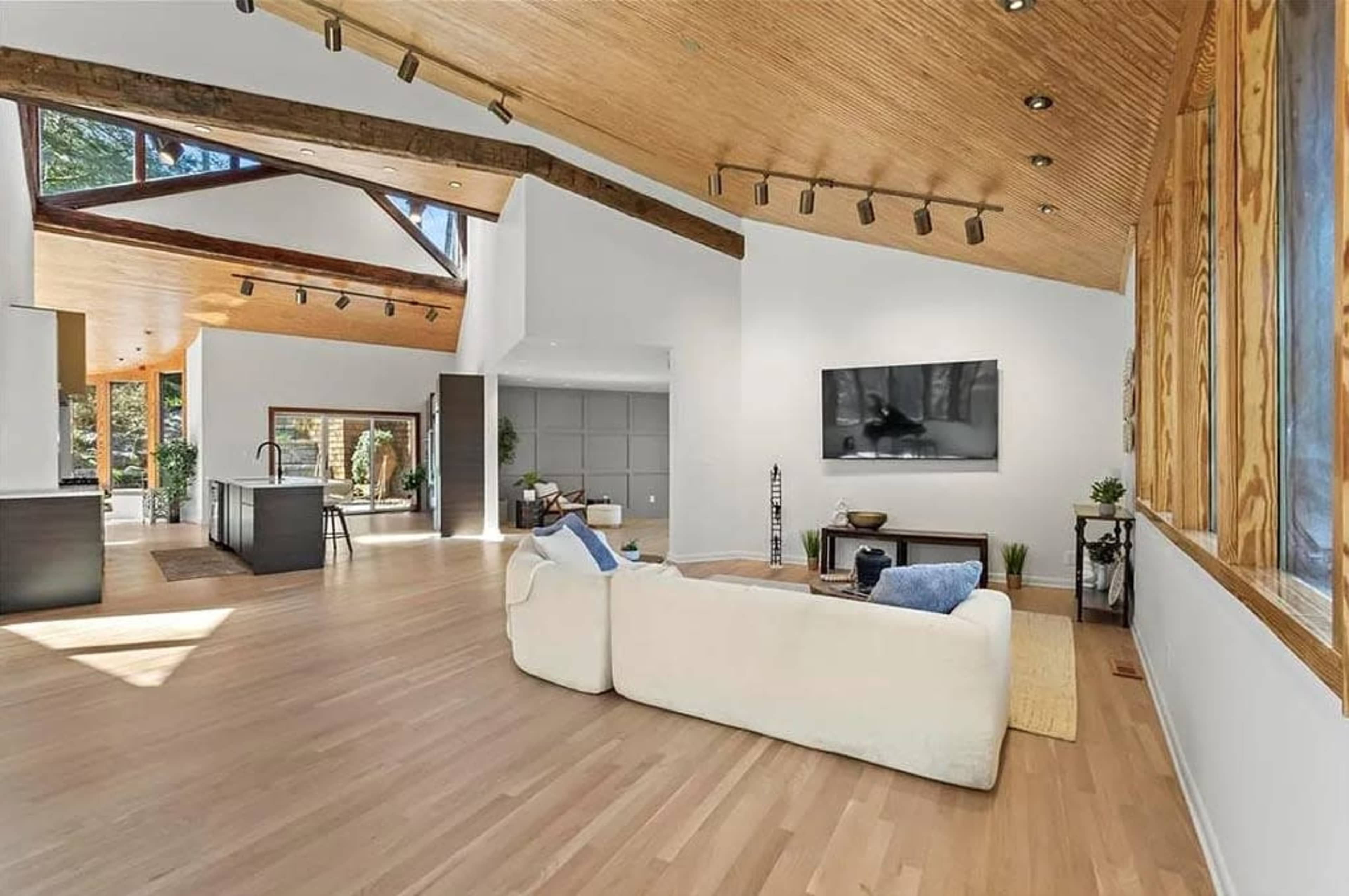 The image shows a modern, open-concept living space featuring a light-colored sofa, wooden beams on a high ceiling, and large windows that illuminate the room.