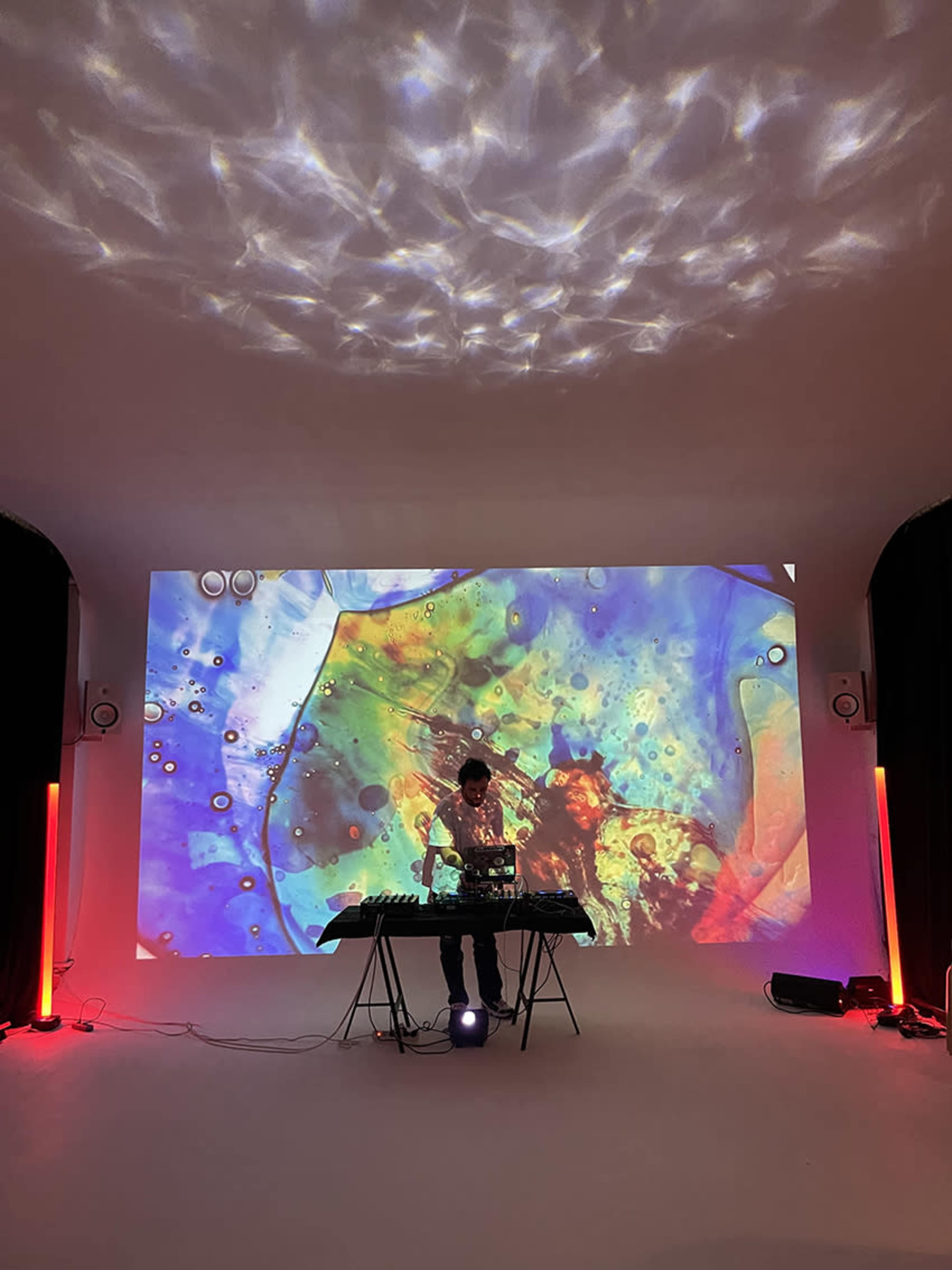 A person stands behind a music equipment setup in a room with colorful abstract visuals projected on a screen behind them.
