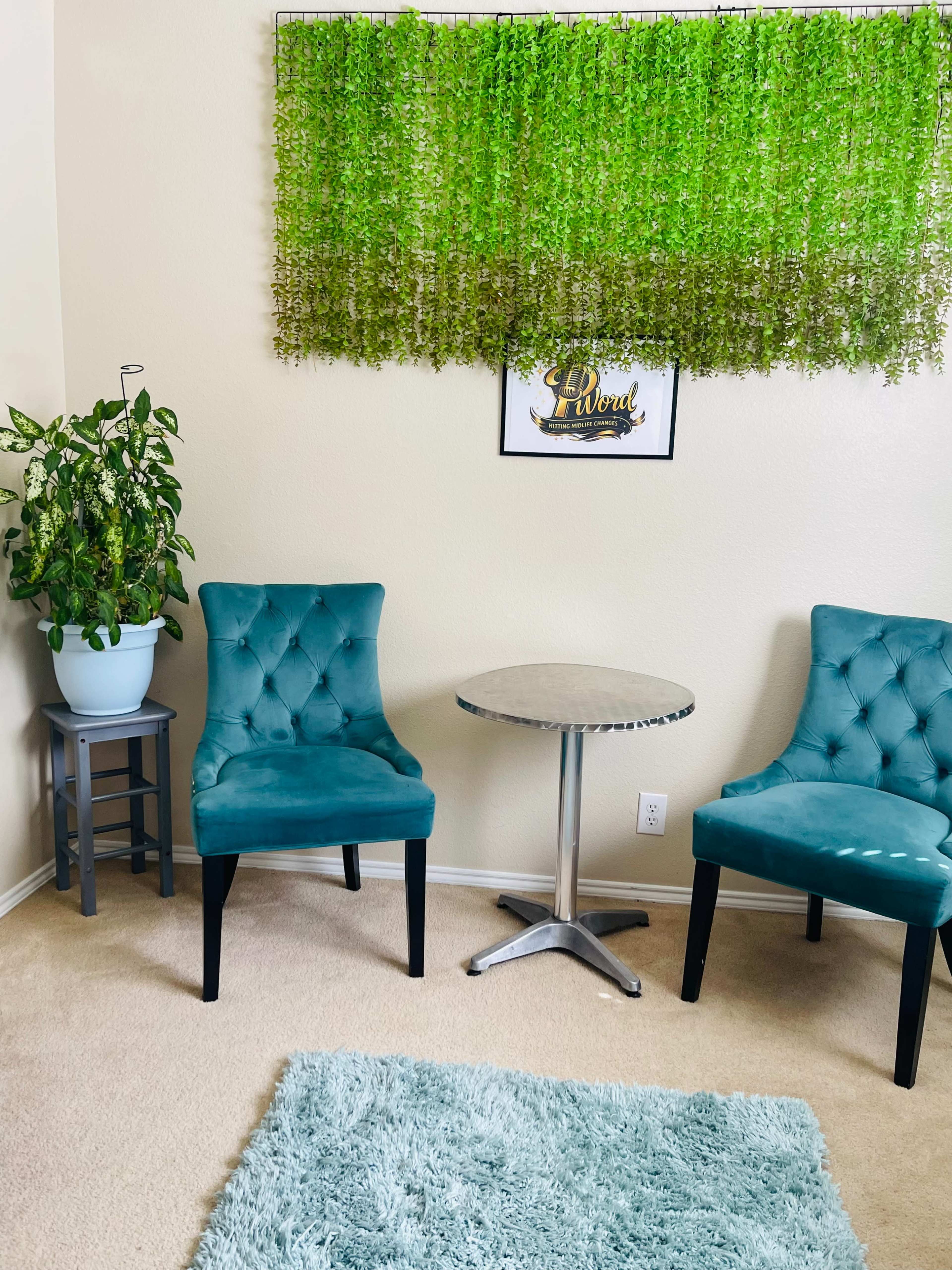 The image shows a small seating area with two teal chairs, a round table, a potted plant, and a decorative green wall hanging above a framed sign.