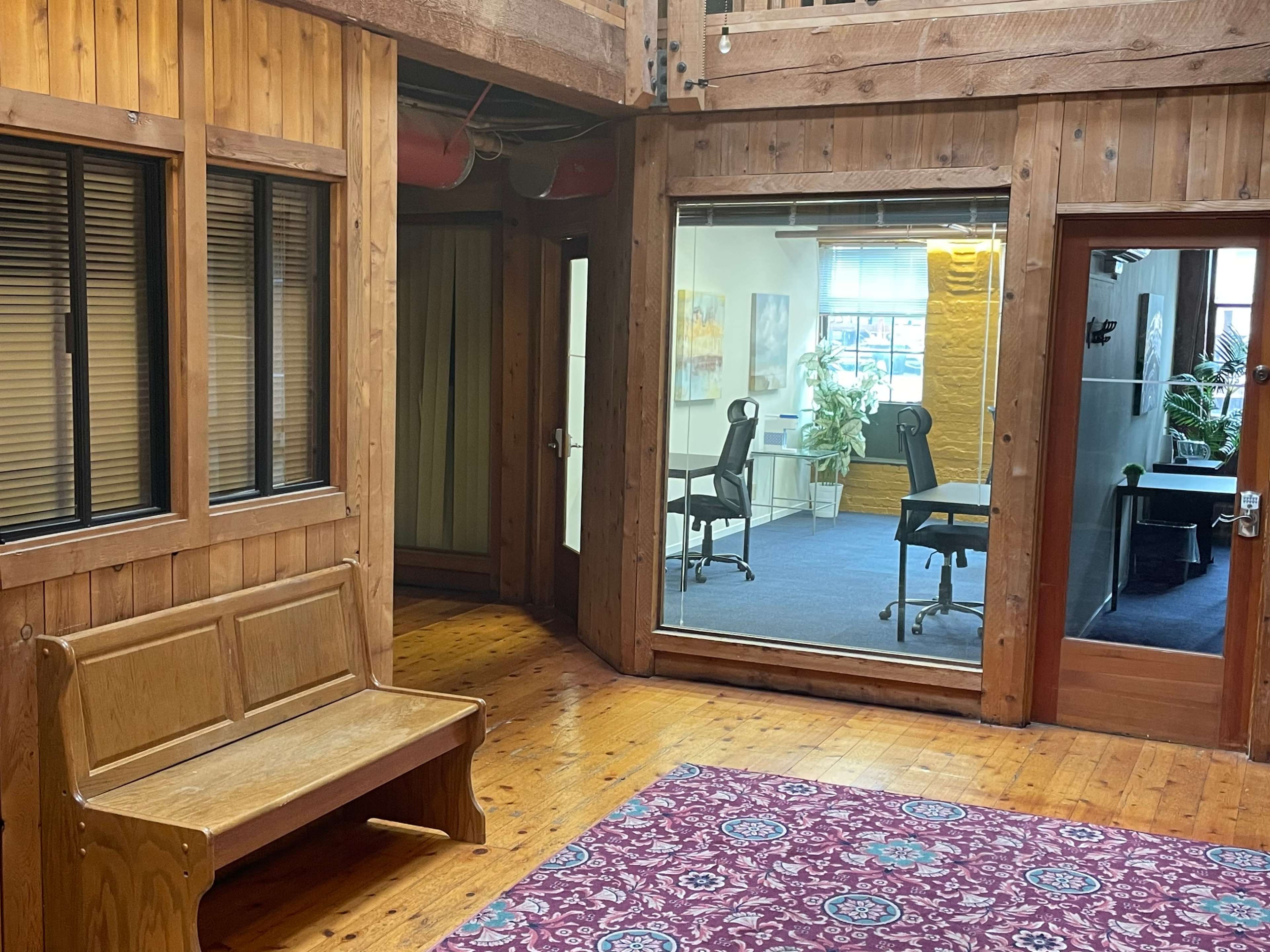 The image shows a wooden interior space with a bench, a patterned rug, and glass-walled offices featuring chairs and plants.